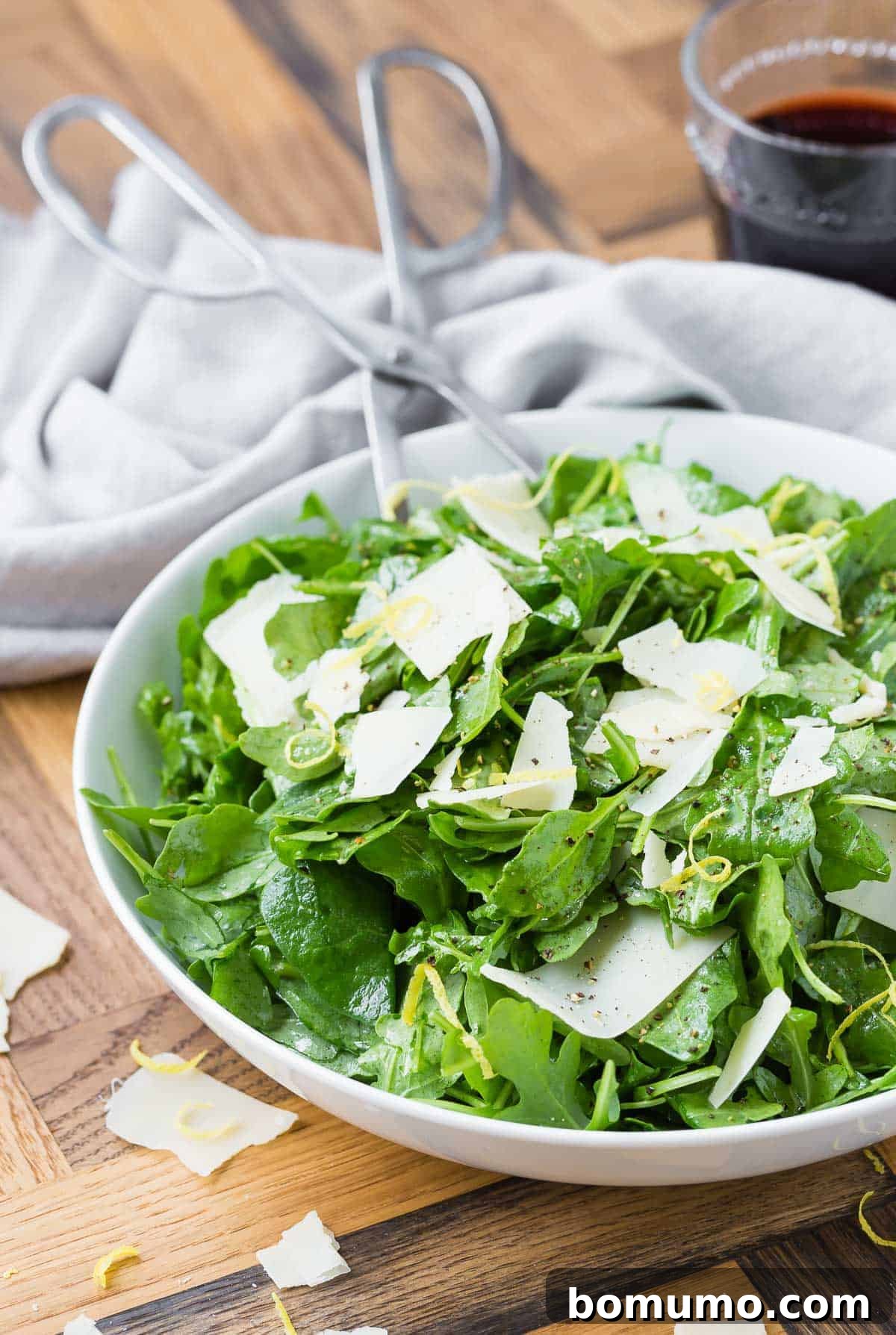 Fresh arugula leaves, lemon zest, and shaved Parmesan cheese arranged in a white bowl on a rustic wooden background.