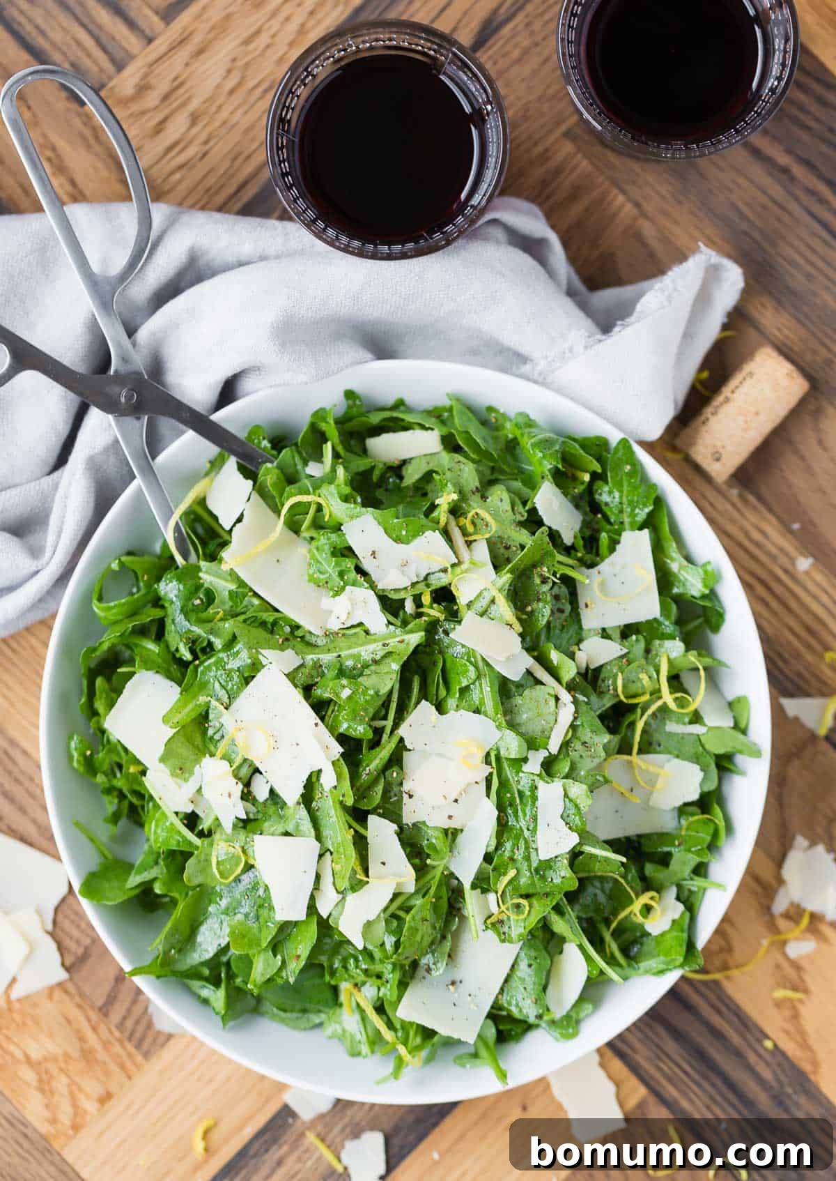 A large ceramic bowl filled with classic arugula salad, garnished with Parmesan shavings and accompanied by silver serving tongs, red wine, and a cork on a wooden surface.