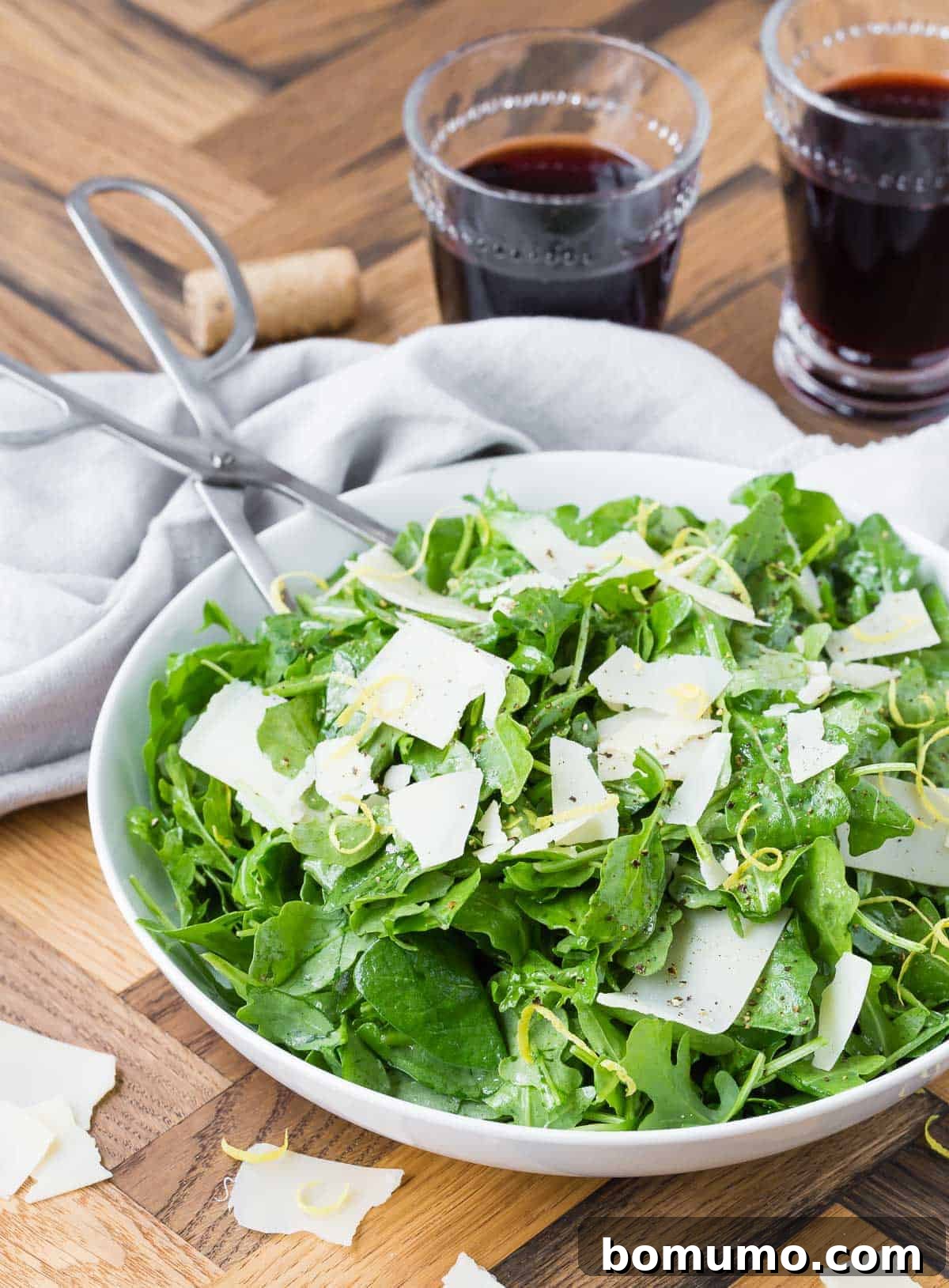 A white bowl showcasing arugula, Parmesan, and lemon zest on a wooden surface, flanked by two glasses of red wine, suggesting a complete dining experience.