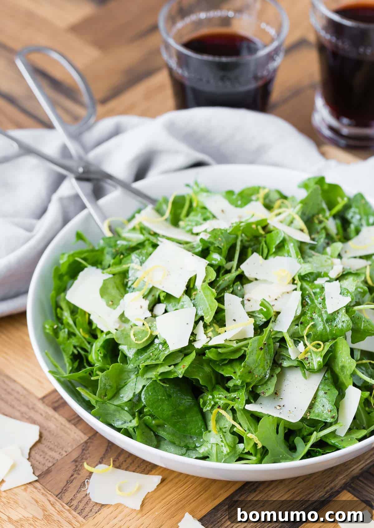 A clean, minimalist shot of a classic arugula salad in a white bowl with silver serving tongs.