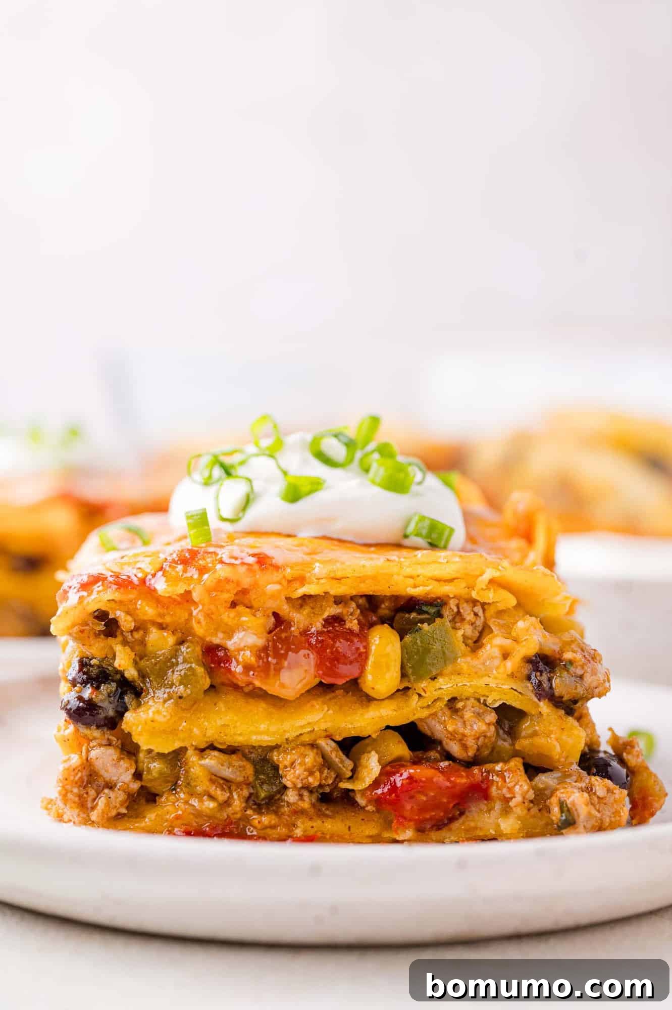 A vibrant slice of layered Mexican casserole on a white plate, showcasing seasoned ground meat, black beans, sweet corn, and colorful vegetables, beautifully crowned with a dollop of sour cream and a sprinkle of fresh chopped green onions.