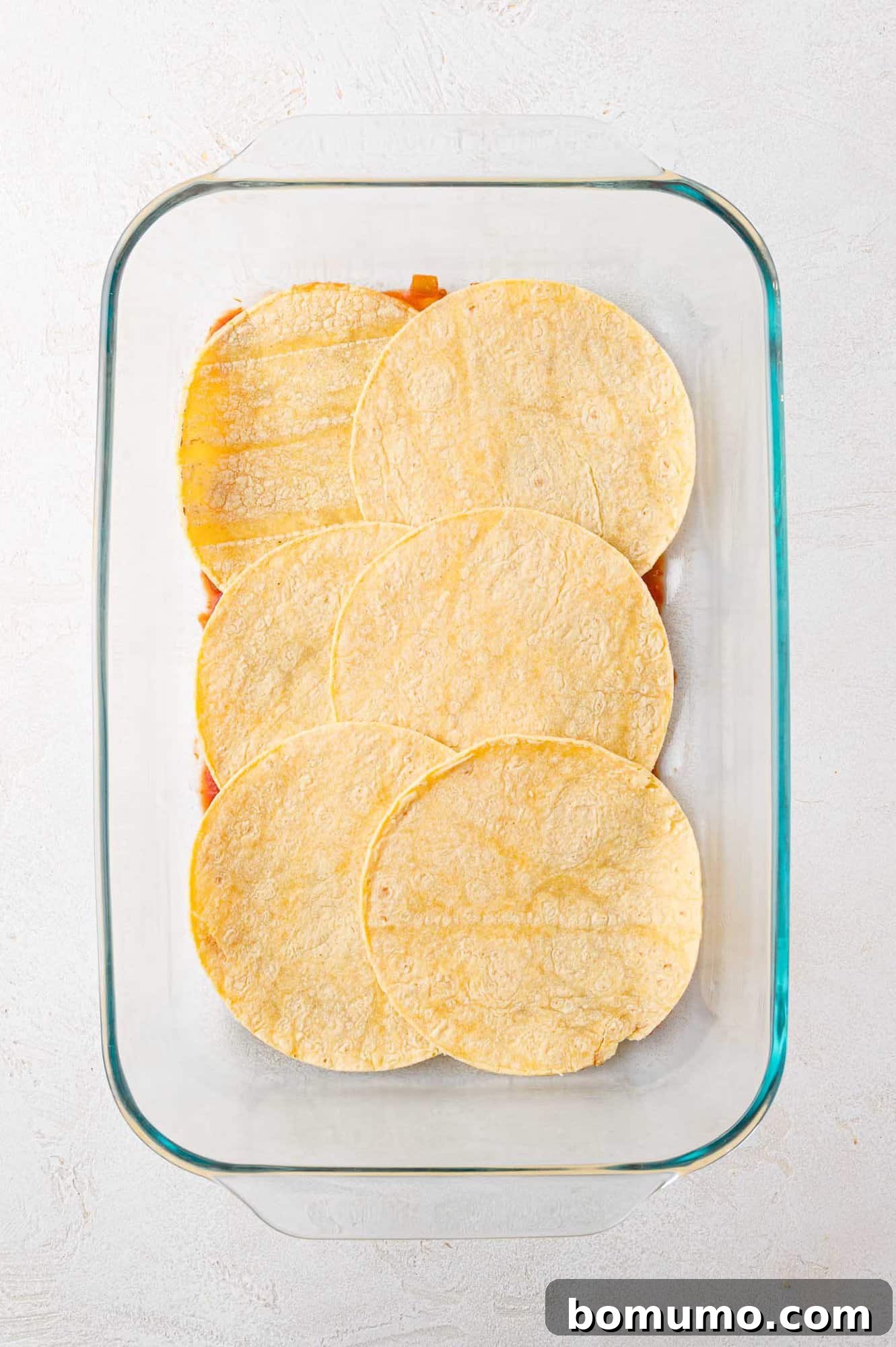 A sturdy glass baking dish showcases six perfectly overlapping corn tortillas, creating a neat, continuous layer that gracefully covers the base ingredients, ready for the next step of assembly.