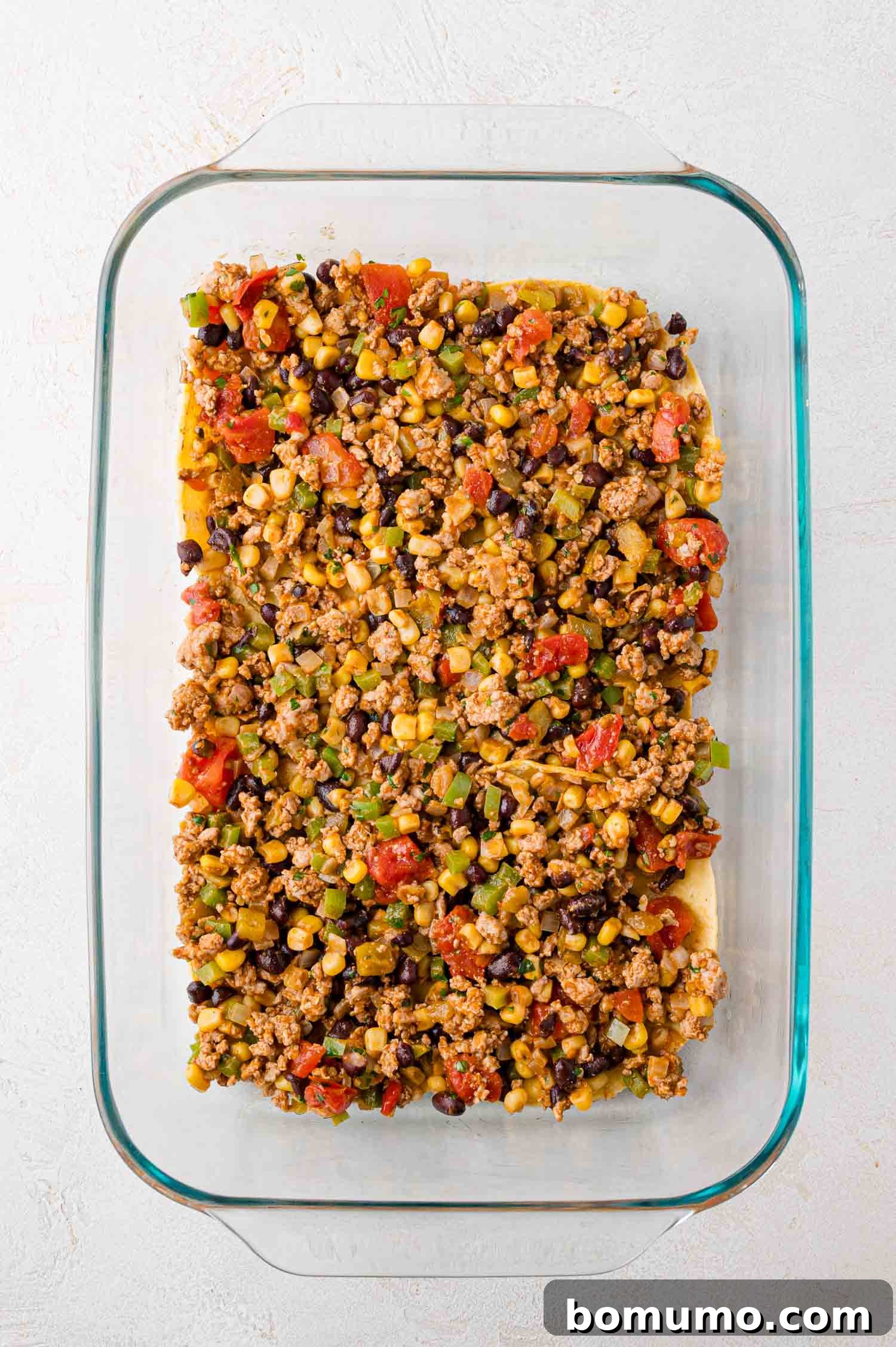 A glass baking dish is generously filled with a vibrant and savory mixture of ground meat, tender black beans, sweet corn, diced tomatoes, and finely chopped vegetables, forming a robust layer on a pristine white surface.