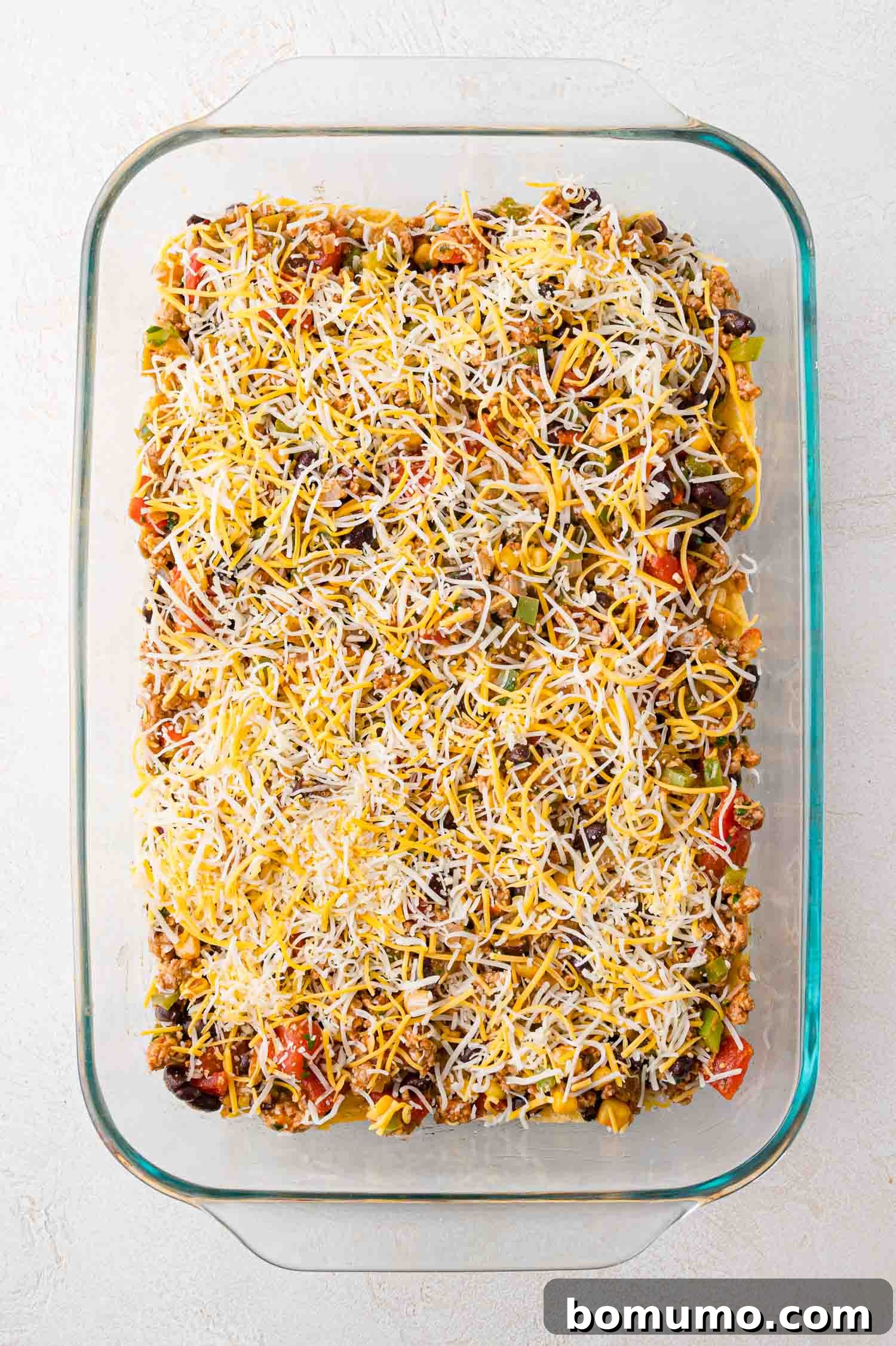A glass baking dish proudly displays an unbaked casserole, rich with layers of seasoned ground meat and vegetables, generously crowned with a blanket of shredded cheese, ready for the oven.