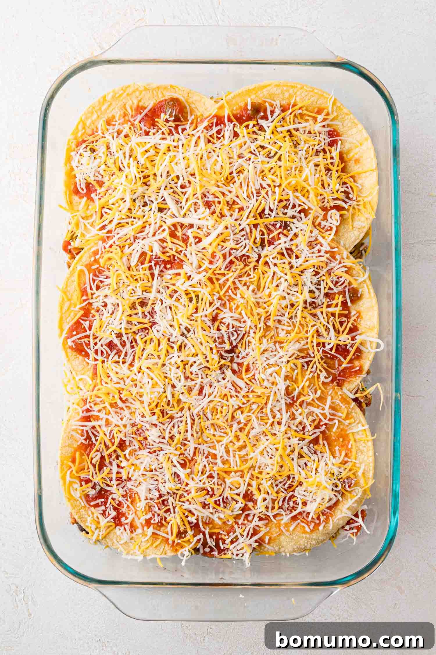 A clear glass baking dish showcases a meticulously layered unbaked casserole, featuring stacked tortillas, vibrant red sauce, and abundant shredded cheese, eagerly awaiting its transformation in the oven.