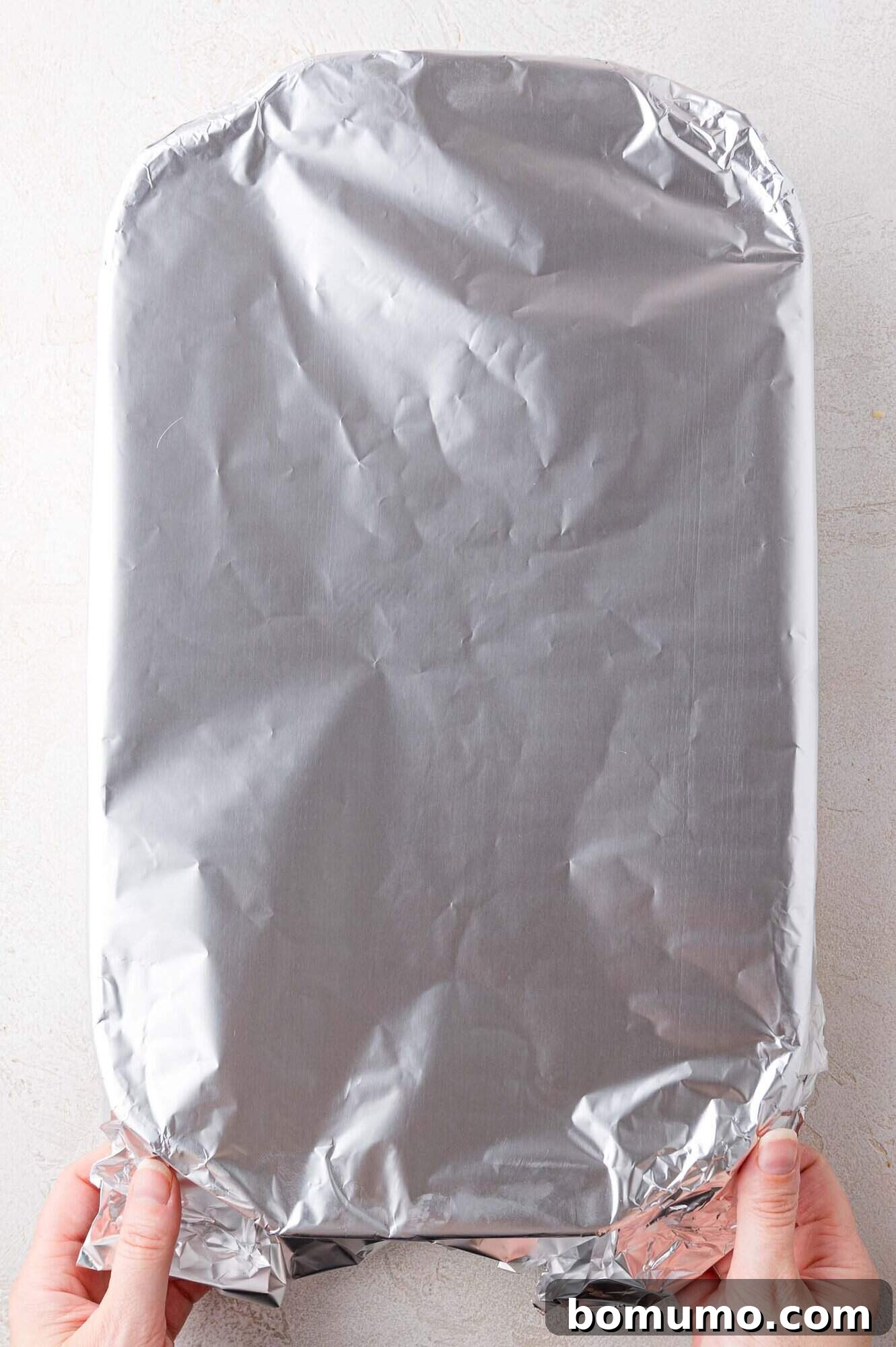 A pair of hands carefully holds a large, rectangular glass baking dish, securely covered with gleaming aluminum foil, positioned on a bright, minimalist surface.