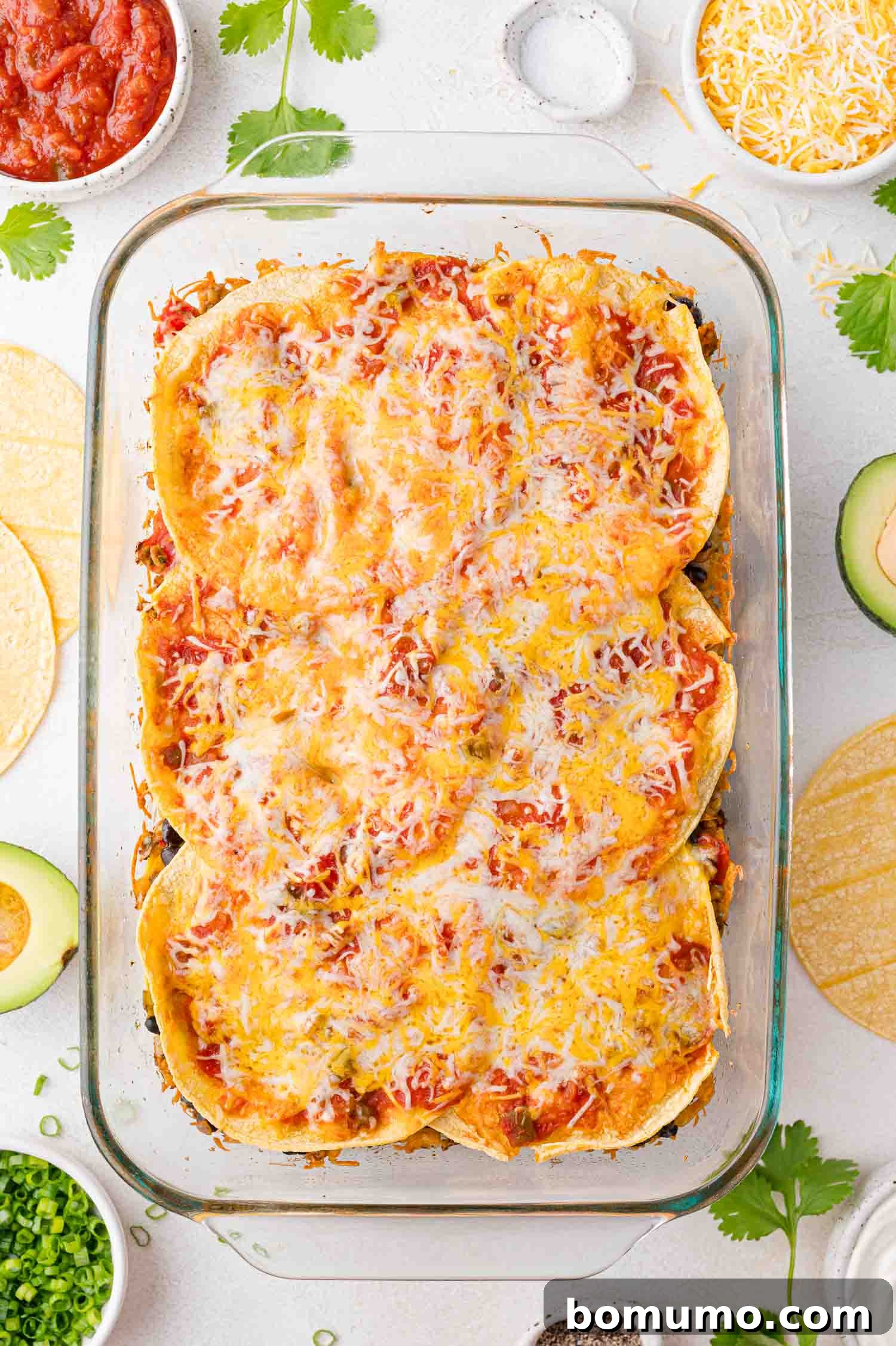 A tempting glass baking dish brimming with a freshly baked, layered Mexican lasagna, topped with an irresistible blanket of perfectly melted cheese. Surrounding it are an assortment of fresh corn tortillas, creamy avocado slices, extra shredded cheese, and small bowls of vibrant salsa and seasonings, all ready for serving.