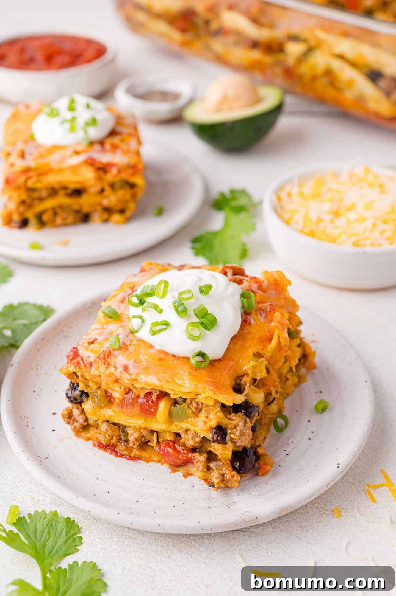A perfect slice of layered Mexican lasagna, artfully arranged on a plate and garnished with cool sour cream and finely chopped green onions, with another serving and a spread of fresh ingredients visible in the softly blurred background.