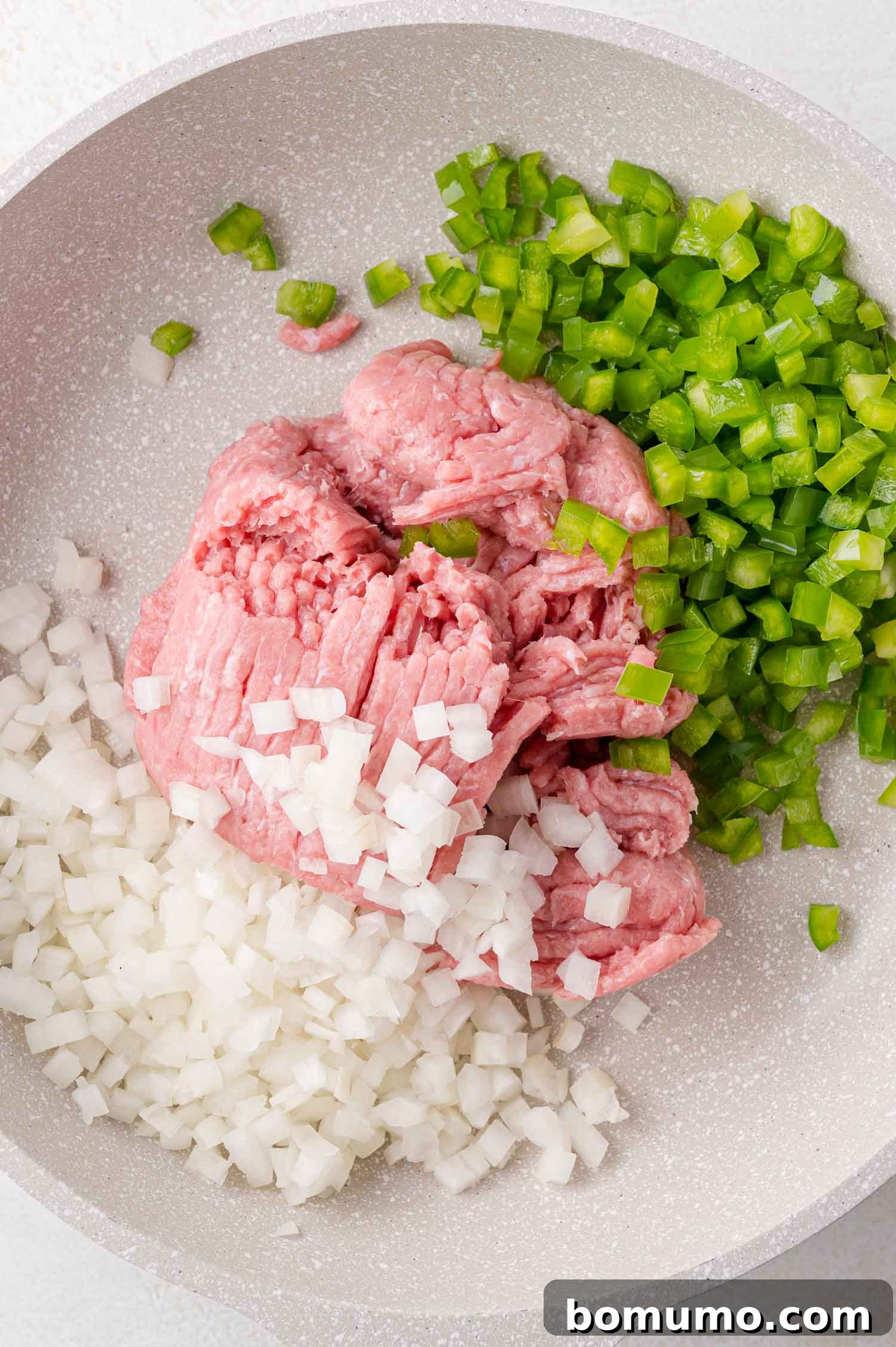 A pristine white frying pan holds raw ground meat, carefully minced white onions, and brightly diced green bell peppers, all poised for cooking to begin.
