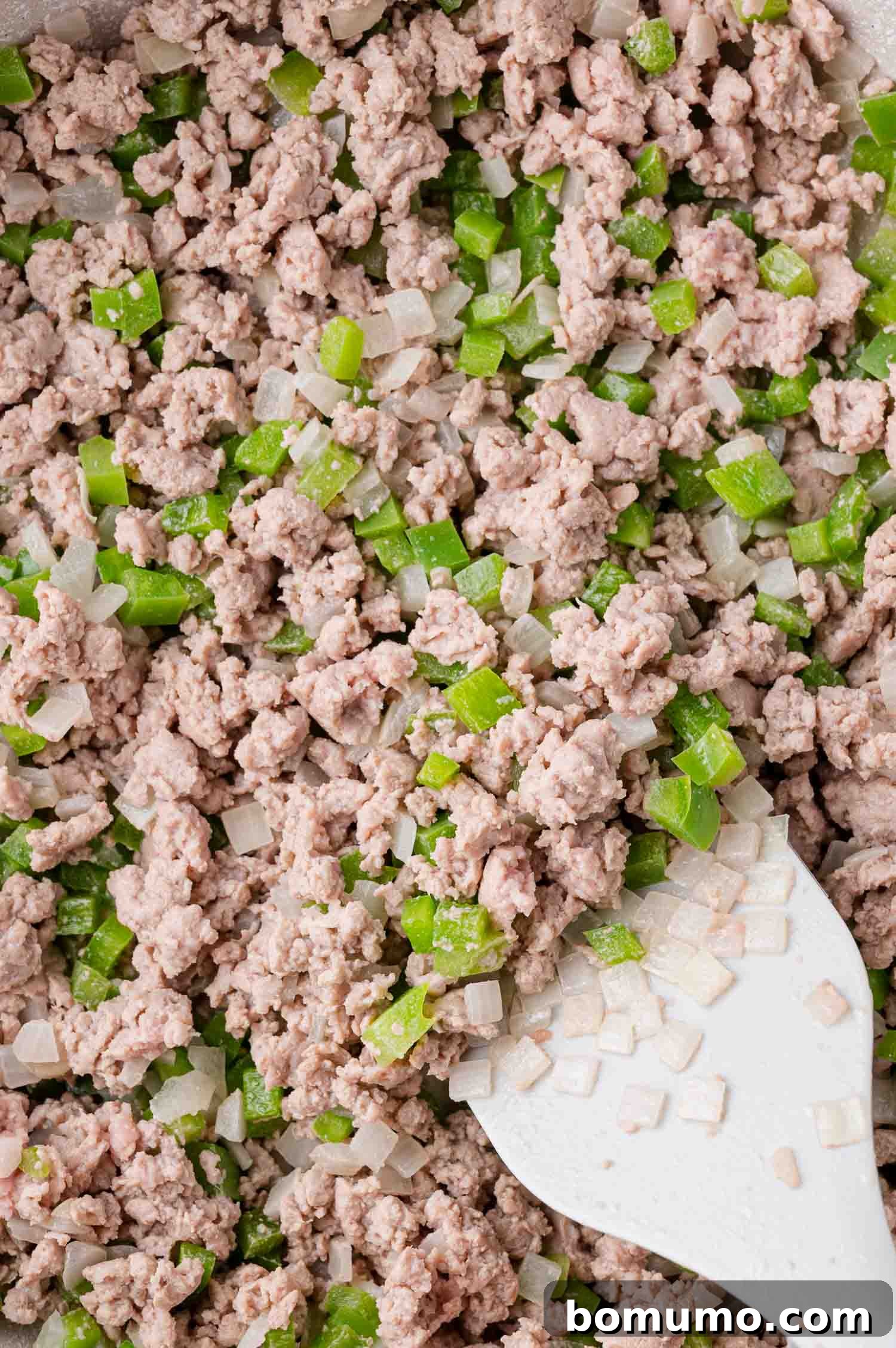 In a pan, perfectly cooked ground meat is mixed with tender diced green bell peppers and onions, actively being stirred with a white spatula to combine the savory ingredients.