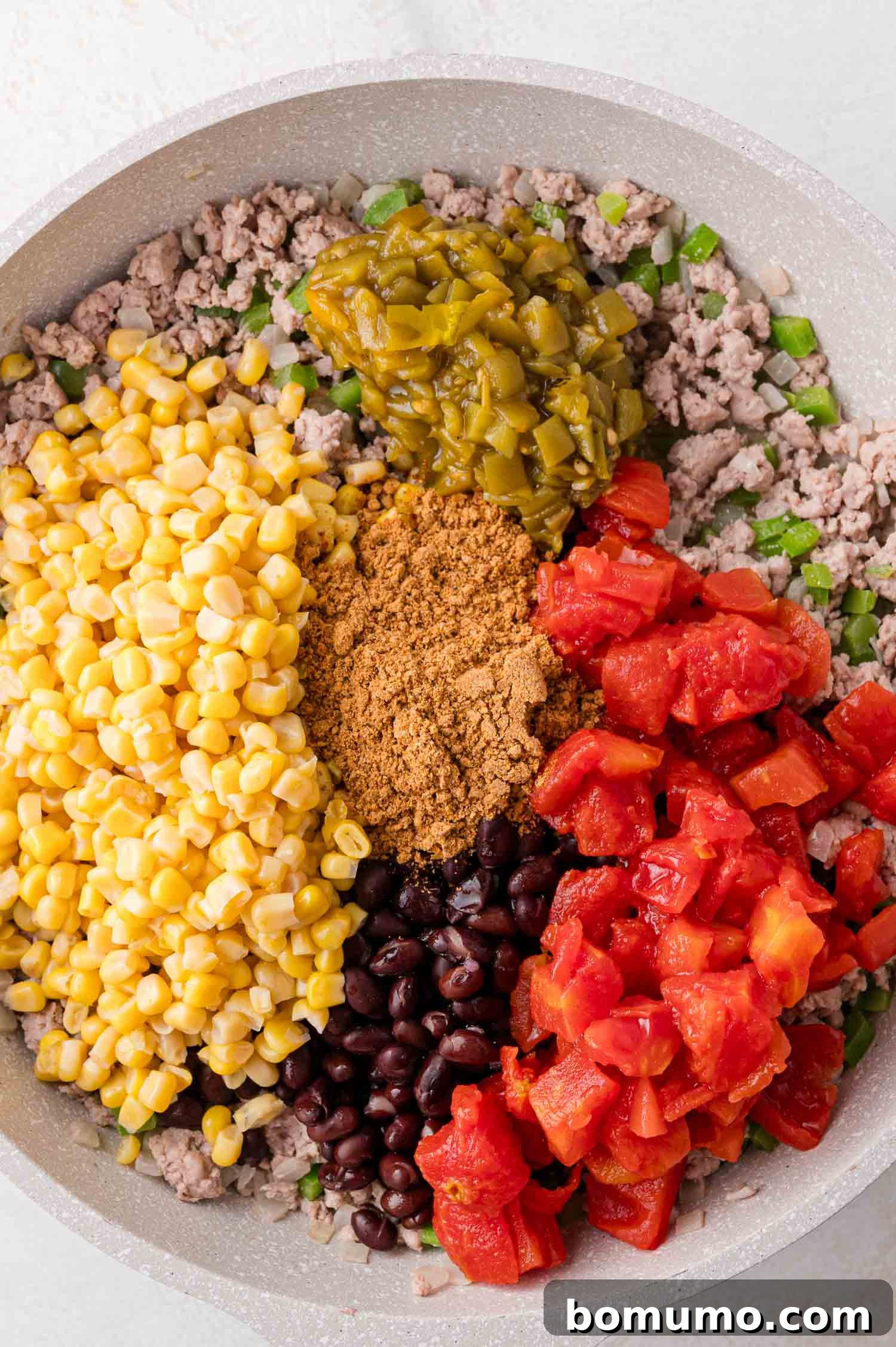 A close-up view of a skillet showcasing the prepared ground meat mixture, alongside distinct sections of corn, black beans, diced tomatoes, green chilies, and taco seasoning, all awaiting combination.
