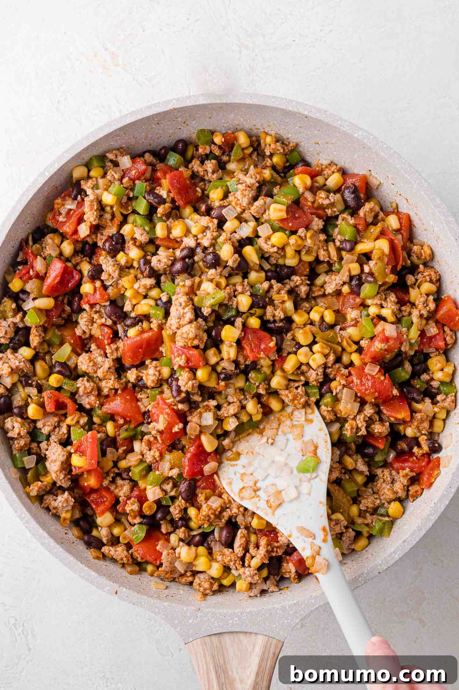 A pan filled with a vibrant mixture of cooked ground meat, sweet corn, savory black beans, diced tomatoes, green peppers, and onions, being expertly stirred with a white spatula.