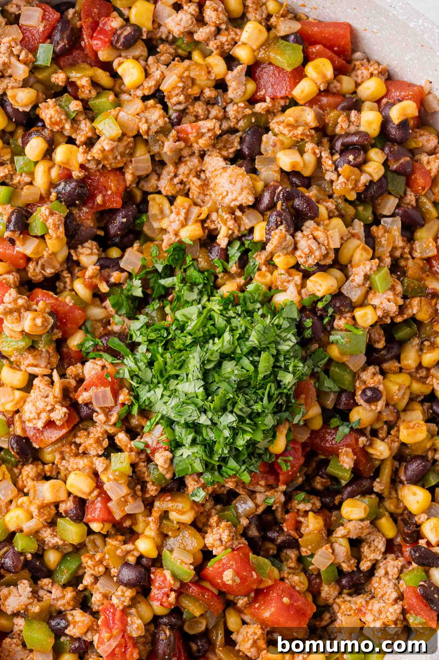 A close-up shot of a rich, cooked mixture containing ground meat, black beans, corn, diced tomatoes, and green bell peppers, beautifully garnished with a generous sprinkle of fresh, chopped cilantro.