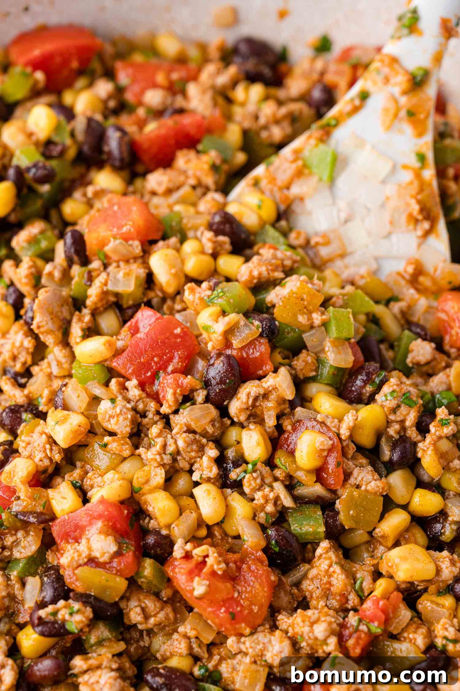 A close-up view of a savory mixture of ground meat, black beans, corn, diced tomatoes, and chopped green peppers, being gently stirred with a white spatula.