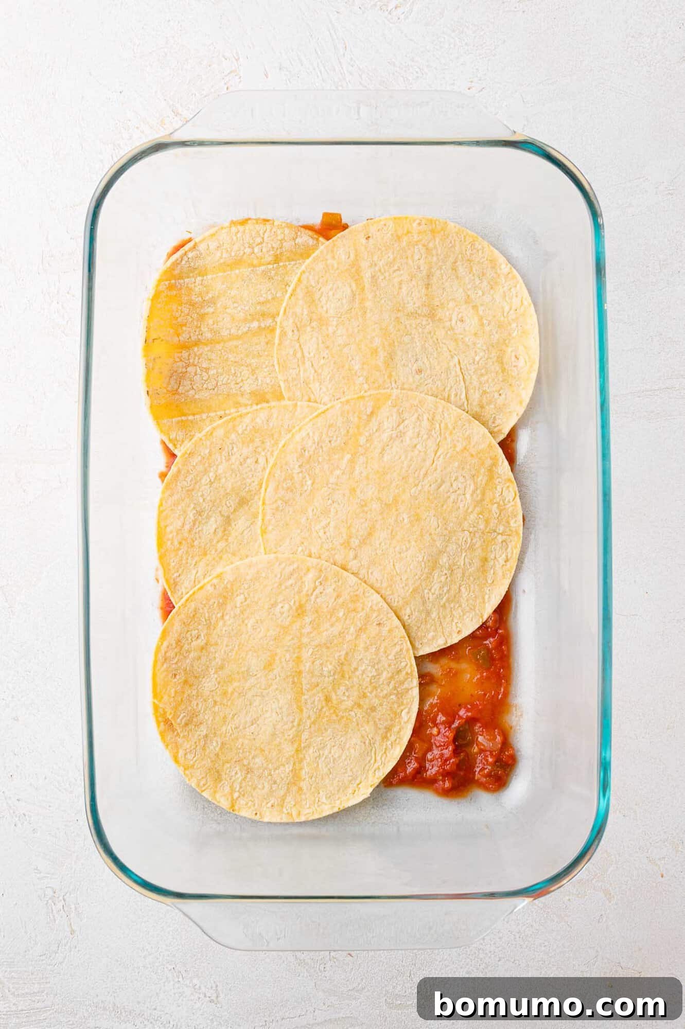 A glistening glass baking dish begins with a vibrant layer of salsa, upon which perfectly round corn tortillas are neatly arranged, creating the foundation for a delicious Mexican lasagna.