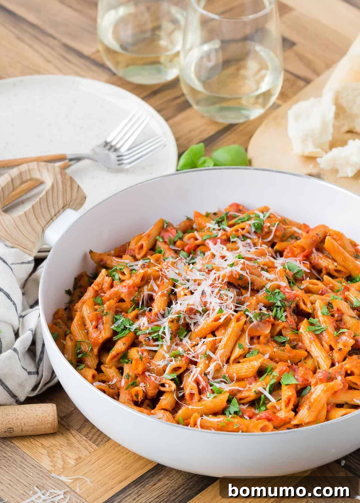 Pasta in a pan, surrounded by two wine glasses, bread, and two plates.