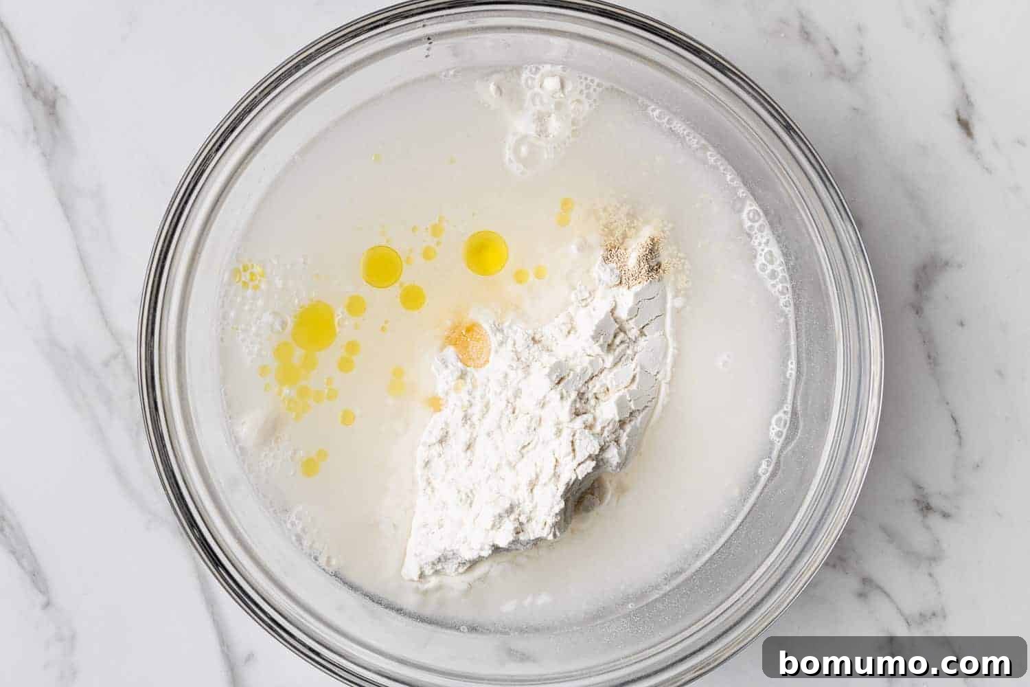 Unmixed bread dough ingredients in a clear glass mixing bowl, ready to be stirred together.
