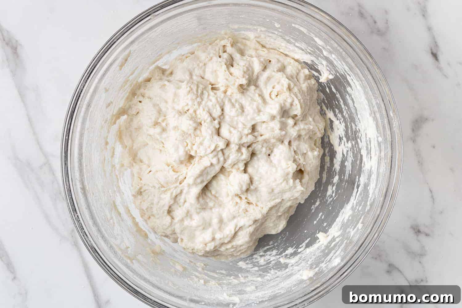 Bread dough, covered tightly with plastic wrap, in a clear glass mixing bowl, after its initial mixing.