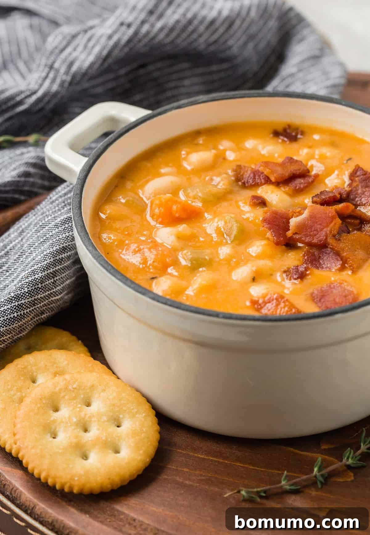 Versatile Bean and Bacon Soup 4 A rustic presentation of bean and bacon soup, served in a white bowl on a wooden tray with a side of crunchy crackers and fresh thyme for garnish.