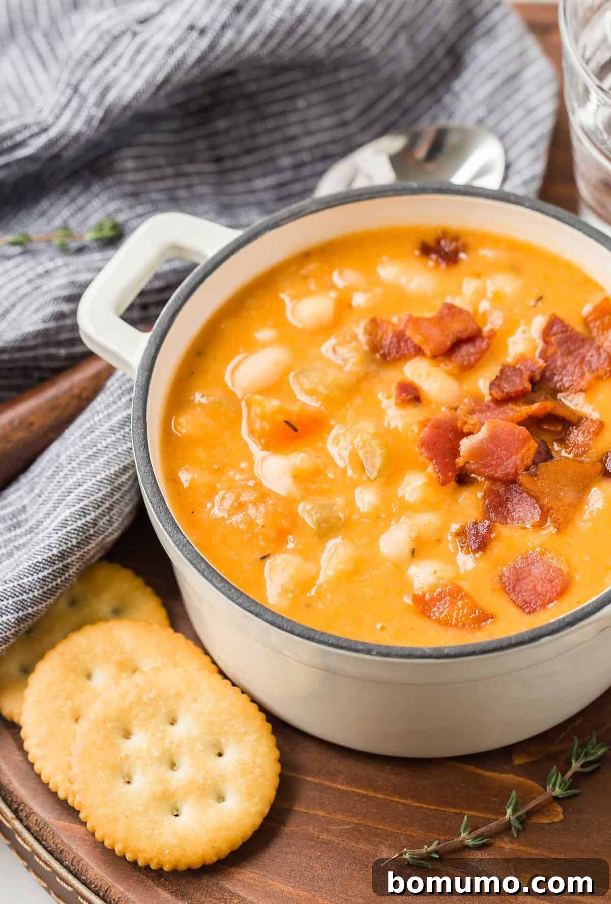 Versatile Bean and Bacon Soup 5 A cozy scene featuring a bowl of homemade bean and bacon soup, adorned with fresh thyme, presented on a light wooden tray with accompanying crackers, inviting a warm, comforting meal.