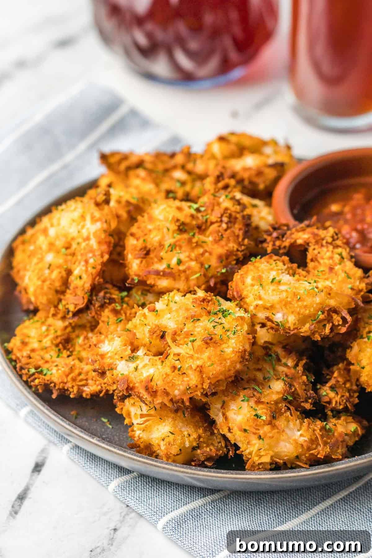 A pile of golden brown and crispy air fryer coconut shrimp on a white plate, served with a small bowl of sweet chili dipping sauce.