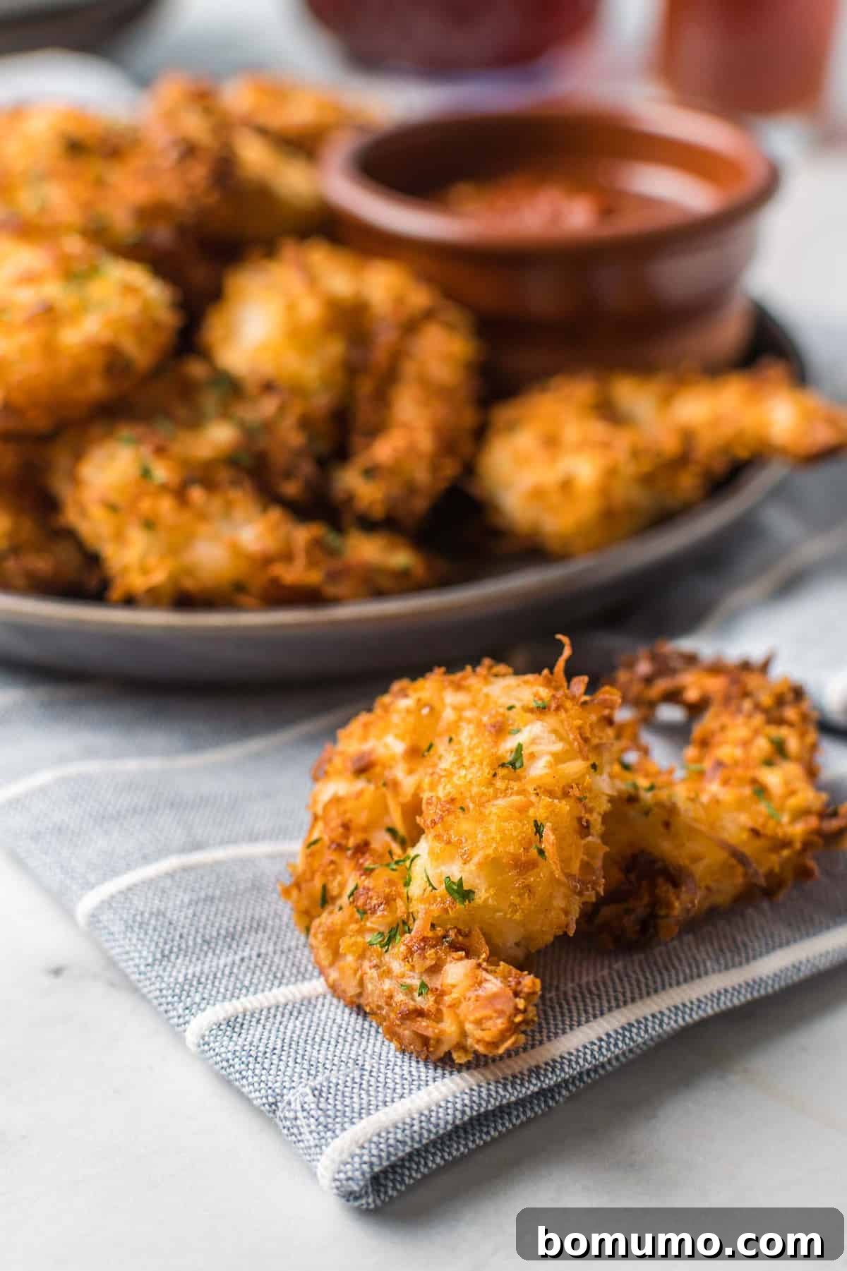 Two perfectly crispy air fryer coconut shrimp are displayed prominently in front of a blurred background of more shrimp on a platter, highlighting their golden brown coating.