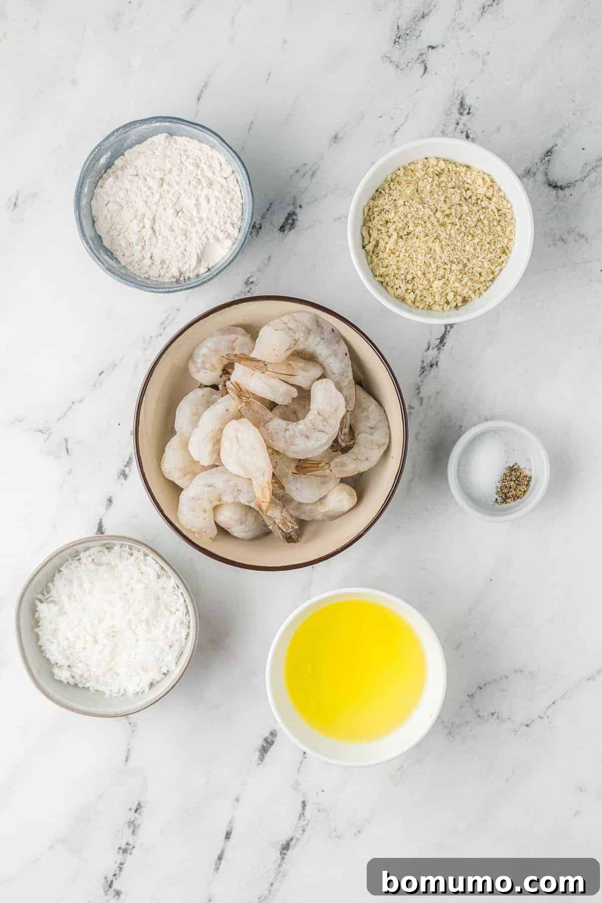 A flat lay photograph showcasing the individual ingredients for air fryer coconut shrimp, including a bowl of shredded coconut, a bowl of beaten egg, a bowl of flour, and a bowl of panko bread crumbs, arranged on a rustic wooden surface.