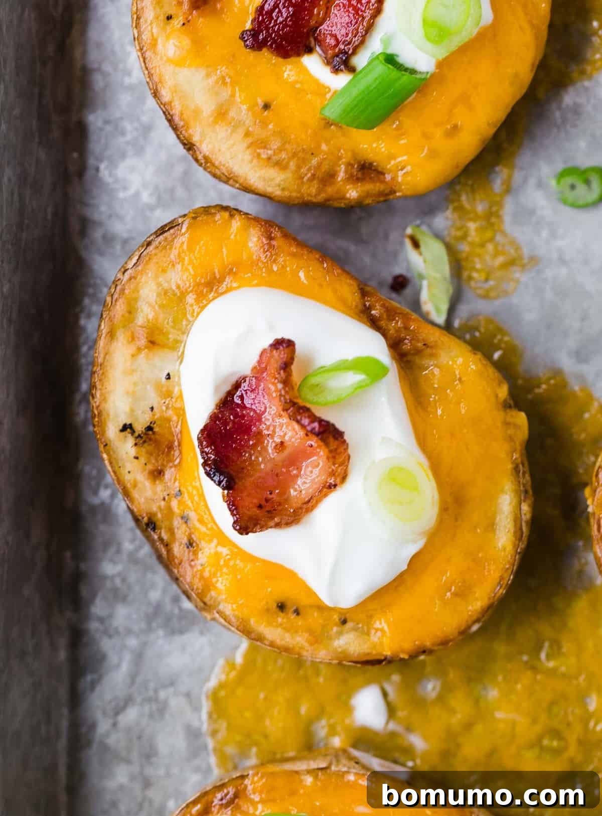 Overhead view of a slice of potato on a tray, covered with cheese, and topped with sour cream, bacon, and green onion.