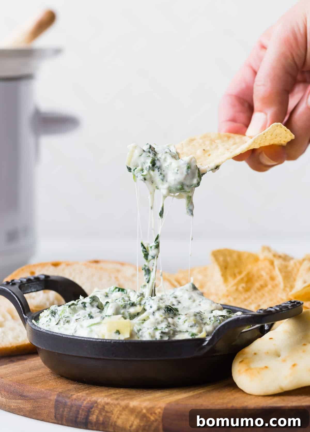 A crispy tortilla chip being gracefully pulled from a bubbling, golden-brown dish of creamy spinach artichoke dip, showcasing its enticing texture and vibrant specks of green.