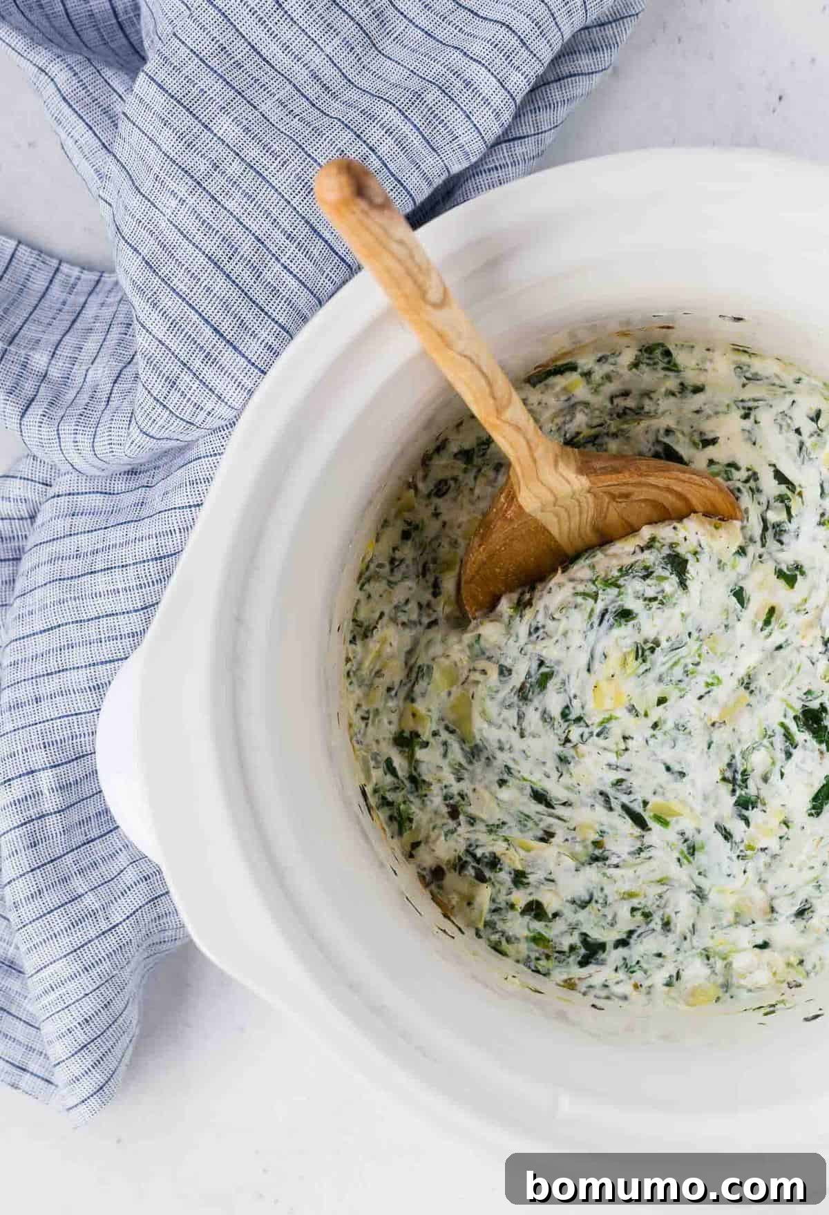 An inviting overhead view of a creamy spinach artichoke dip simmering in a white slow cooker, its cheesy surface gently bubbling and ready to be served.