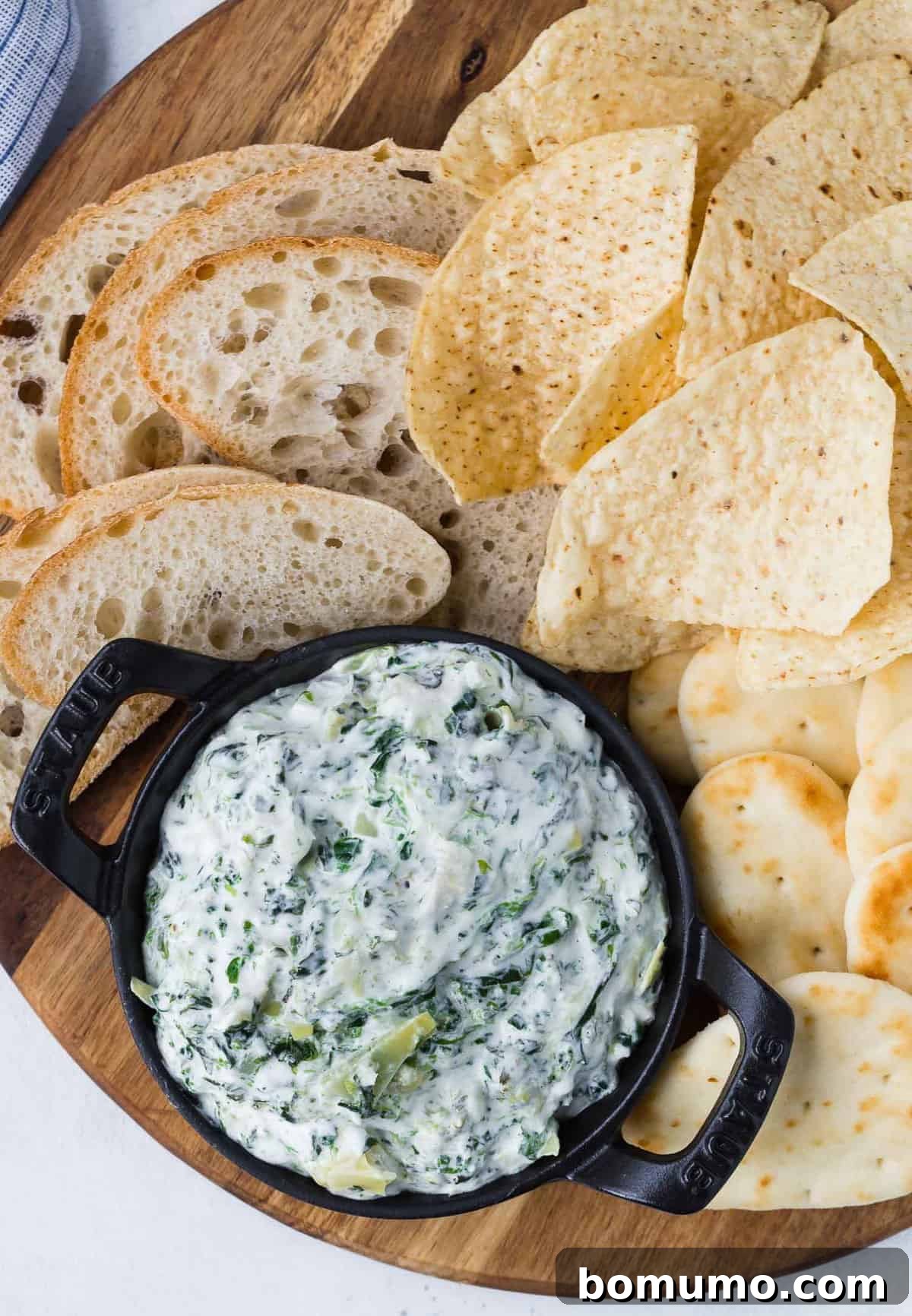 An overhead shot showcasing a small black dish filled with rich spinach artichoke dip, artfully surrounded by an assortment of golden tortilla chips and crusty bread slices, inviting immediate indulgence.
