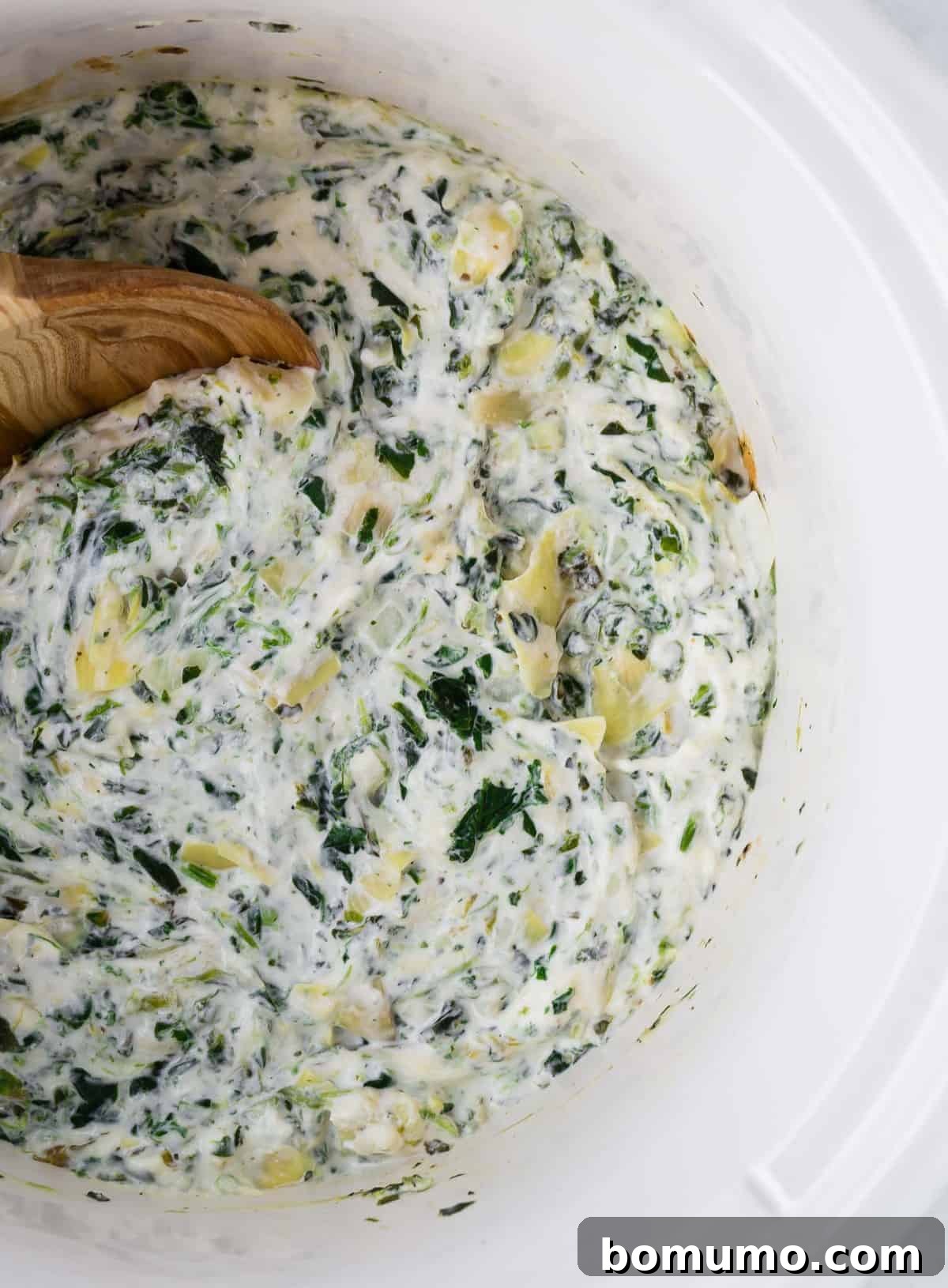 An inviting overhead shot of creamy spinach artichoke dip, featuring visible artichoke hearts and lush green spinach, nestled in a white slow cooker alongside a rustic wooden spoon.