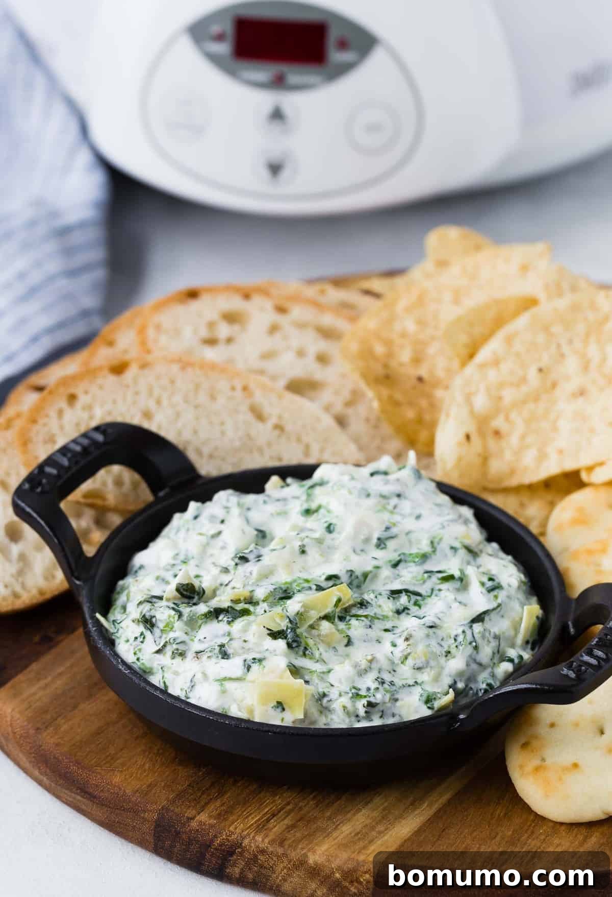 A small, elegant black bowl of creamy spinach artichoke dip, artfully placed on a rustic wooden board, accompanied by an array of crunchy chips and slices of artisan bread, perfect for sharing.