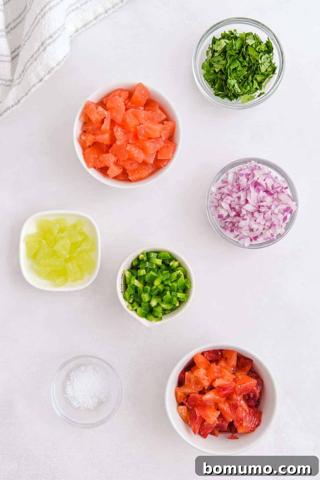 Overhead view of several small bowls holding individual ingredients for citrus salsa: finely diced red onions, jalapeños, segmented grapefruit, oranges, fresh cilantro, and lime wedges.