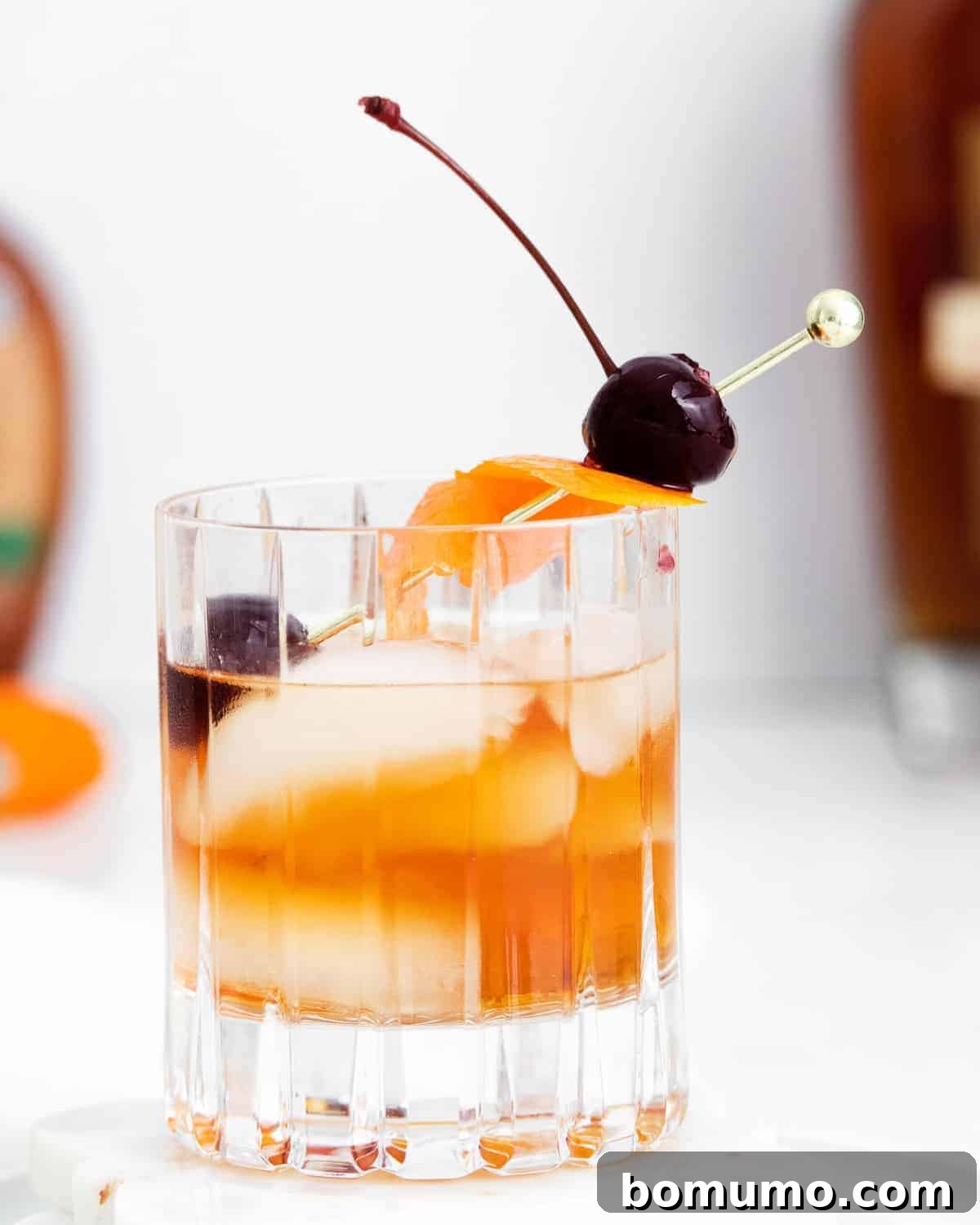 A short cocktail glass filled with ice, an orange peel, a maraschino cherry, and golden whiskey, ready to be enjoyed.