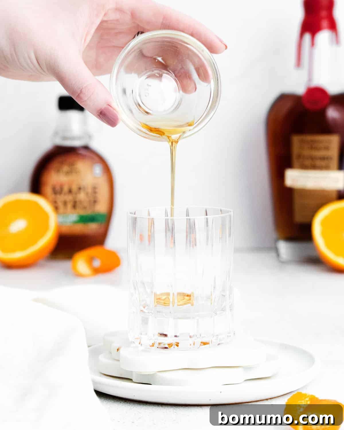A hand gently pours pure maple syrup from a small glass bowl into an empty rocks glass. In the soft-focus background, bottles of whiskey and maple syrup, along with sliced oranges, suggest a well-stocked bar.