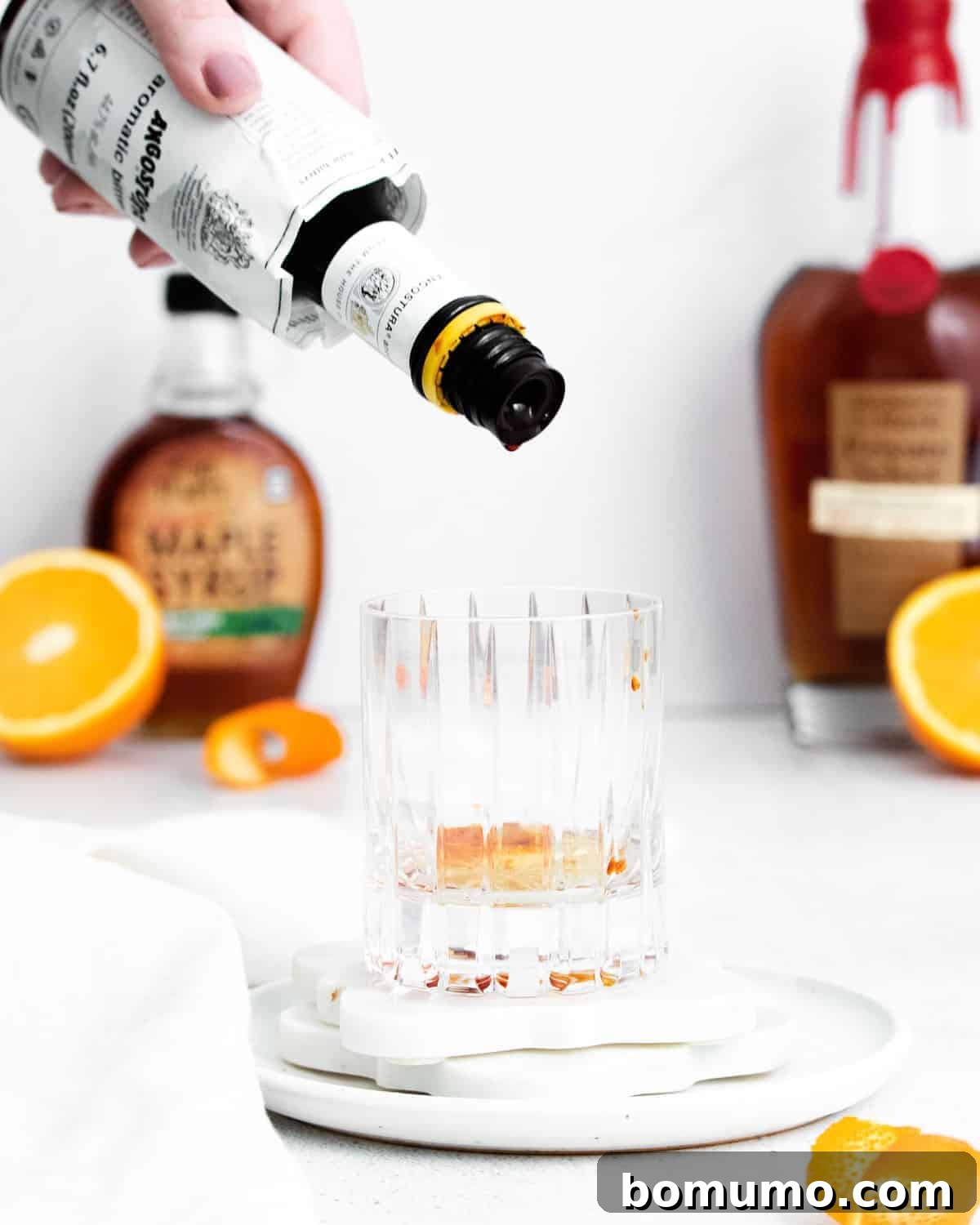 A skilled hand carefully adds dashes of aromatic bitters into an empty glass. The background features various bottles of spirits and syrups, along with vibrant orange slices, indicating the preparation of a sophisticated cocktail.