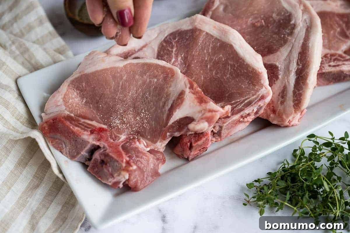 A chef seasoning thick-cut pork chops with salt and freshly ground black pepper on a cutting board, preparing them for searing.