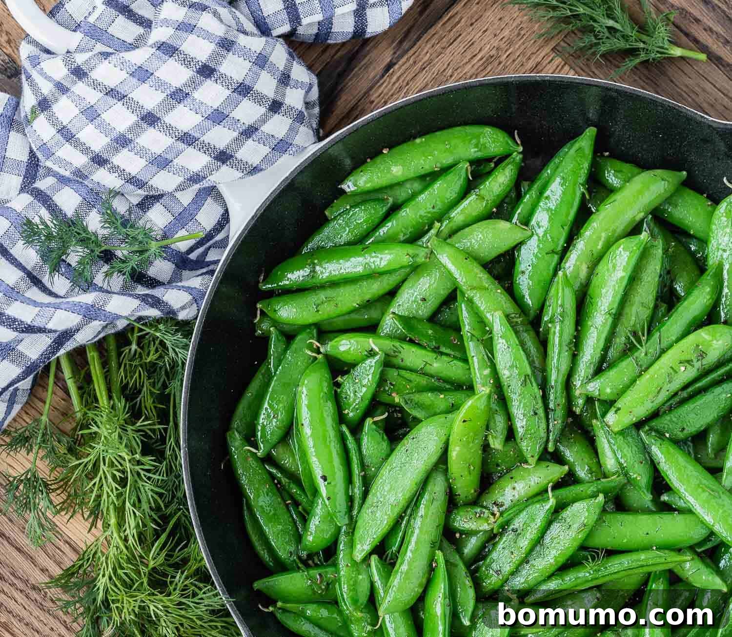 Cooked snap peas with dill in a pan on a wooden surface with a blue and white linen.