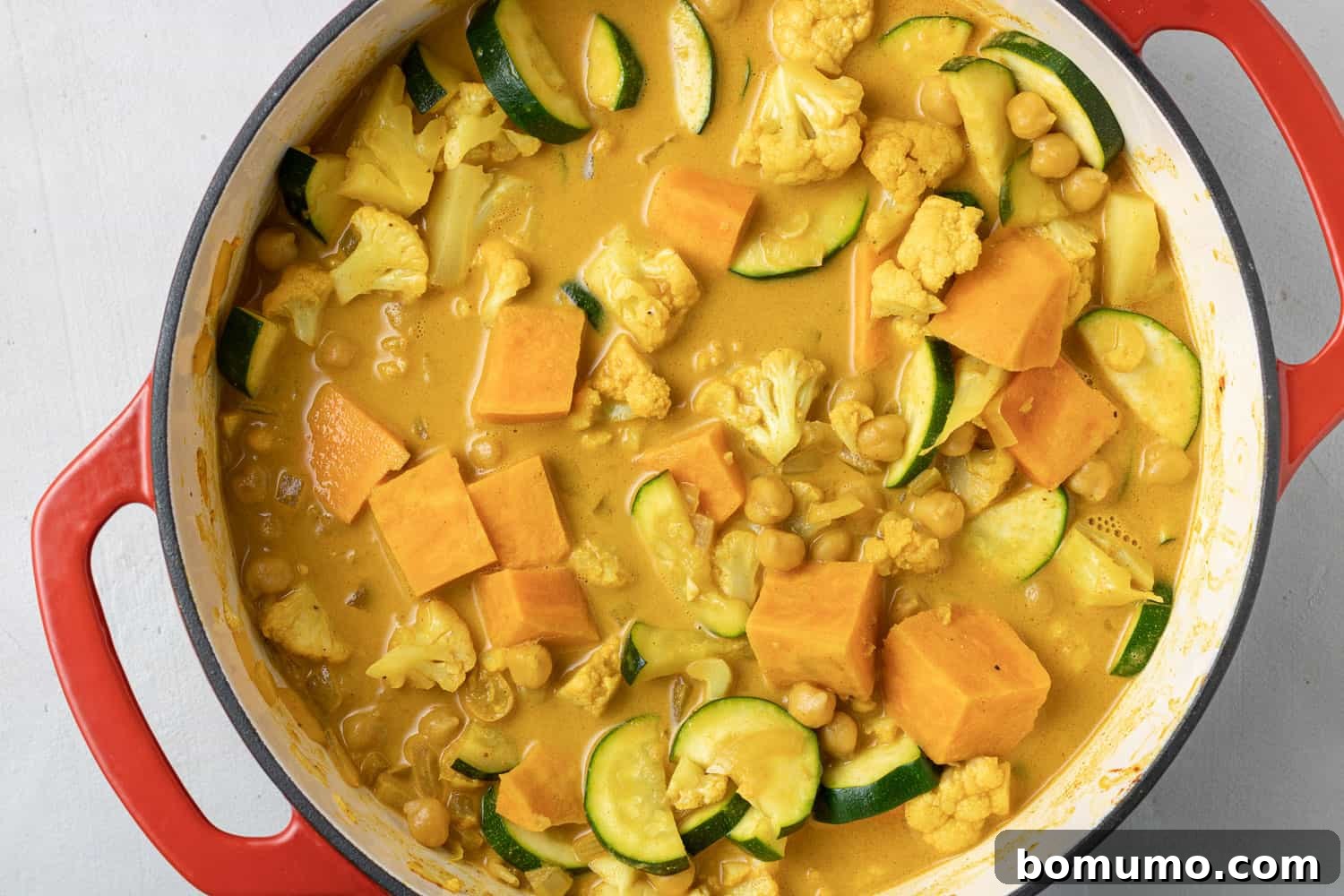 An enticing view of uncooked vegetarian curry ingredients, including vibrant vegetables and rich liquids, arranged in a deep red Dutch oven on a stylish gray background, ready to simmer.