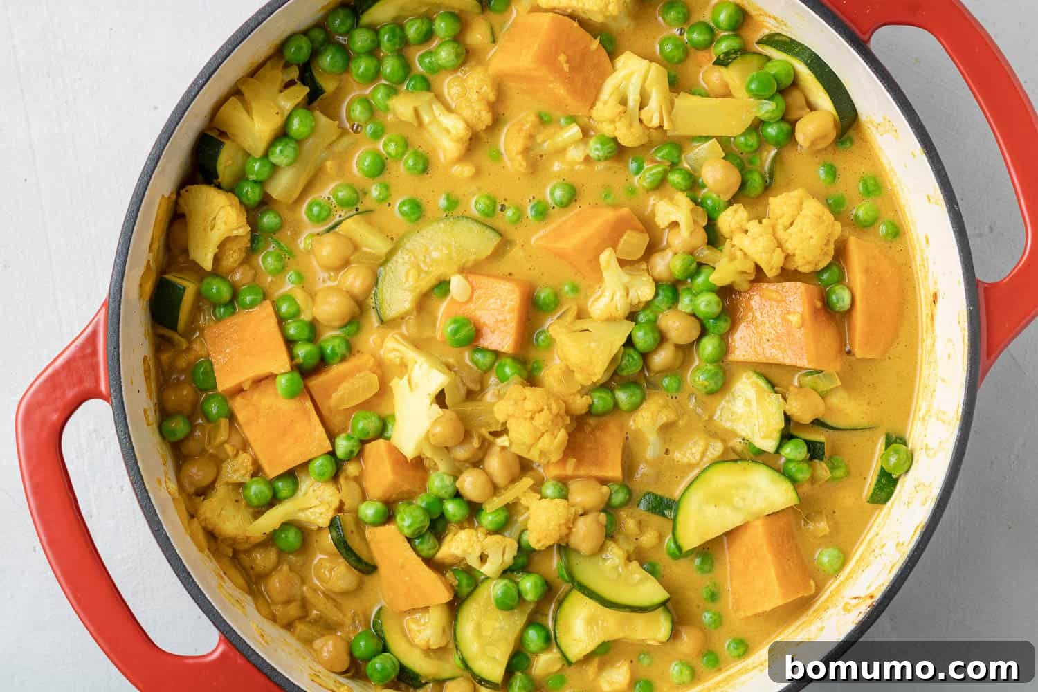 Vibrant green peas being added to a rich, yellow-orange curry in a striking red Dutch oven, illustrating the final steps of preparation.