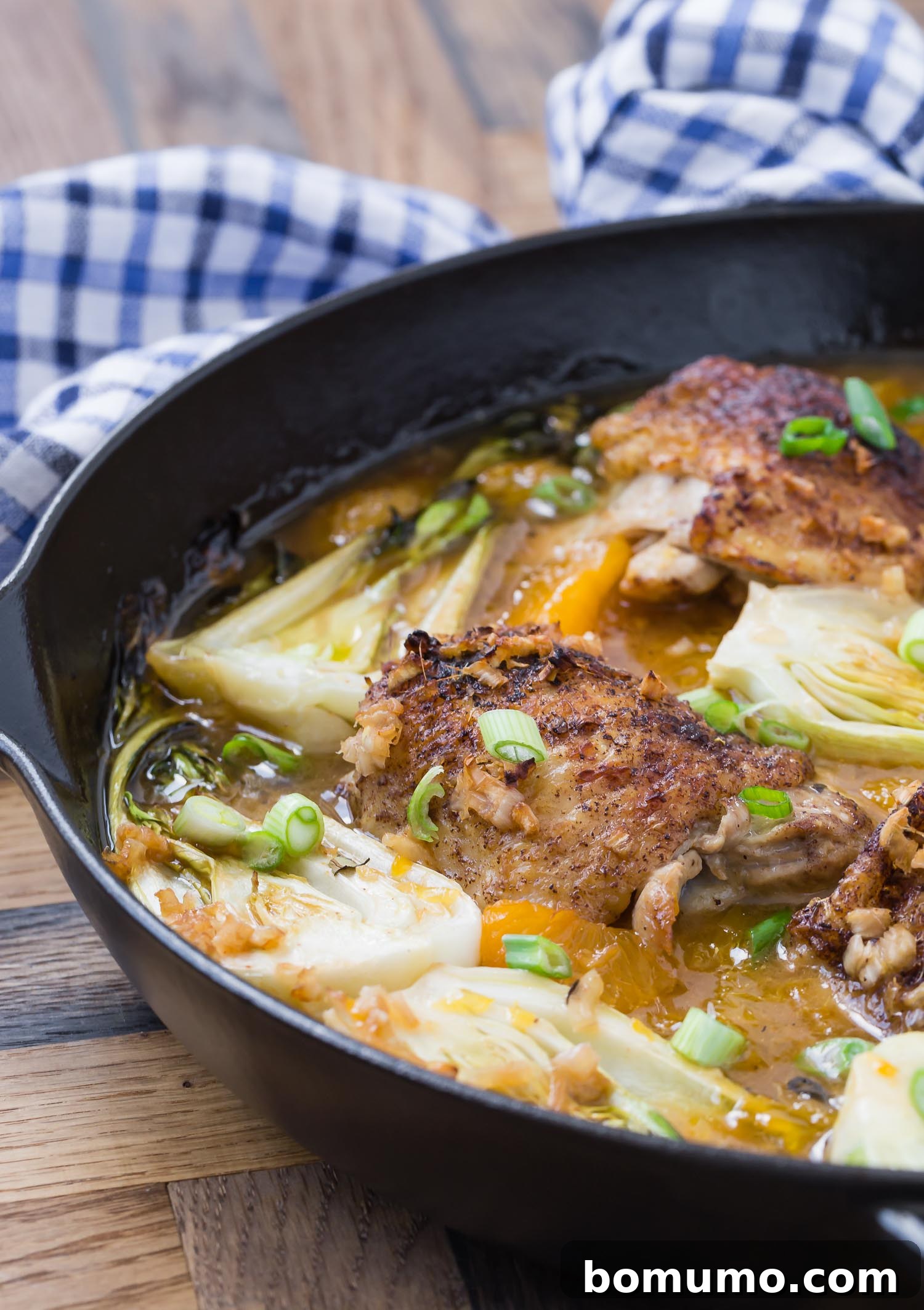 Black skillet with orange chicken thighs and bok choy, on a wooden surface.