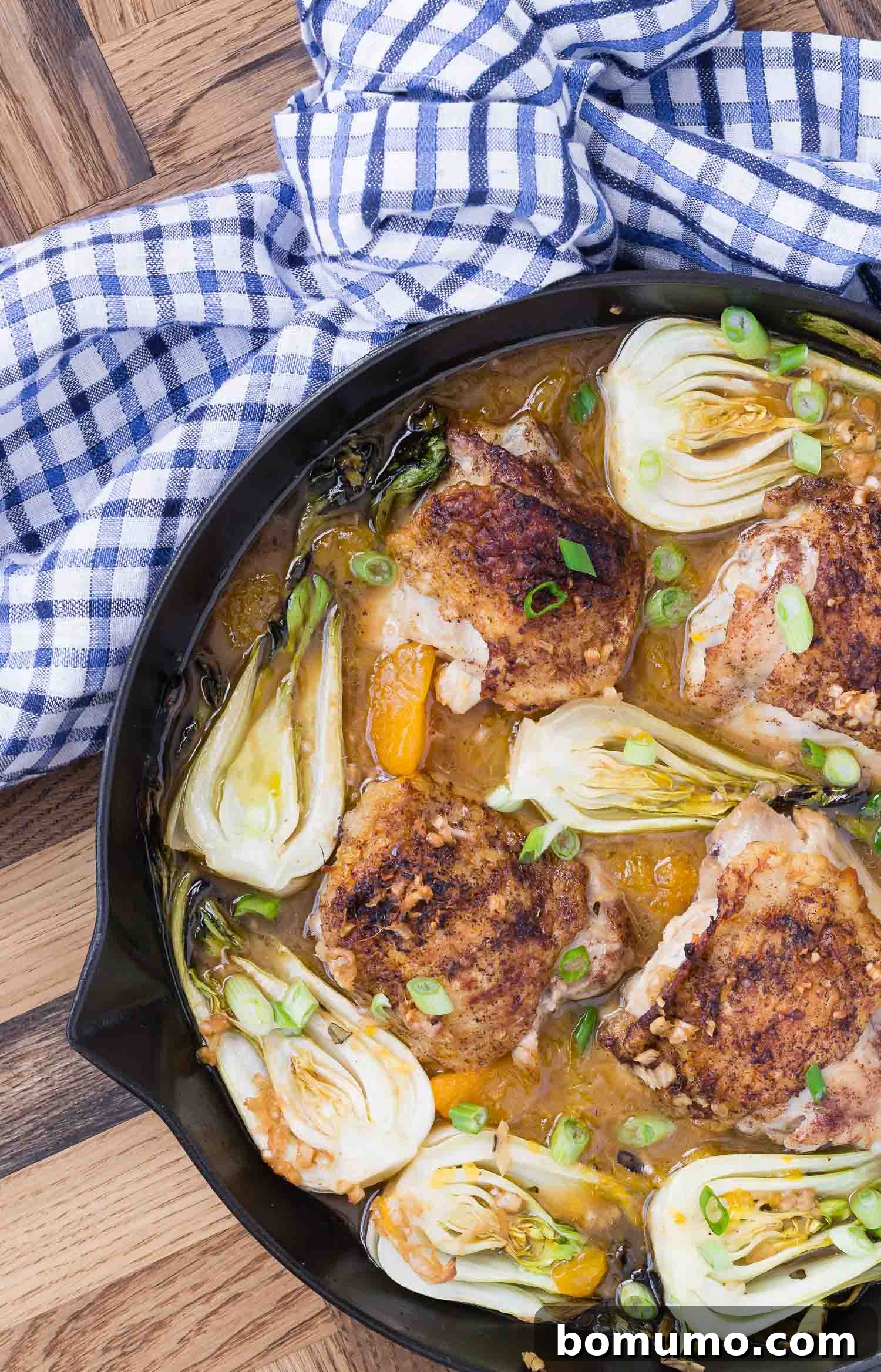 Overhead view of chicken thighs and baby bok choy in a black pan with a blue and white linen.
