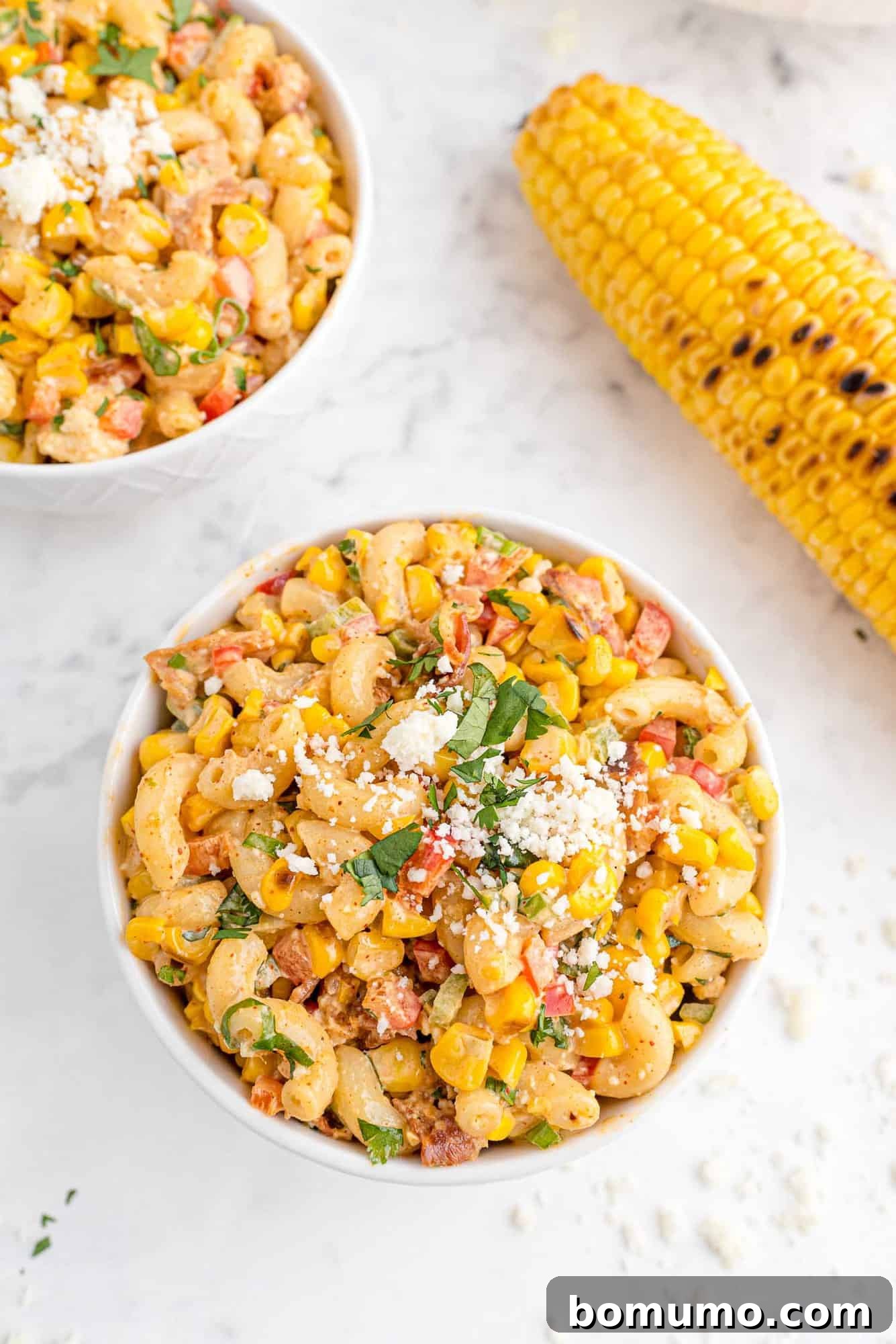 A beautifully composed image of the pasta salad in a bowl, artfully garnished with fresh cilantro and delicate white cheese, with a charred corn cob resting beside it.