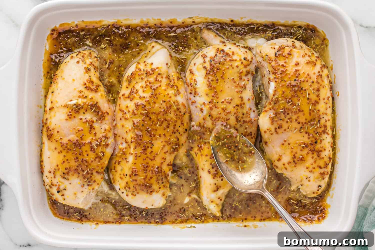 Rich honey mustard sauce being delicately spooned over golden-brown chicken in a baking dish.