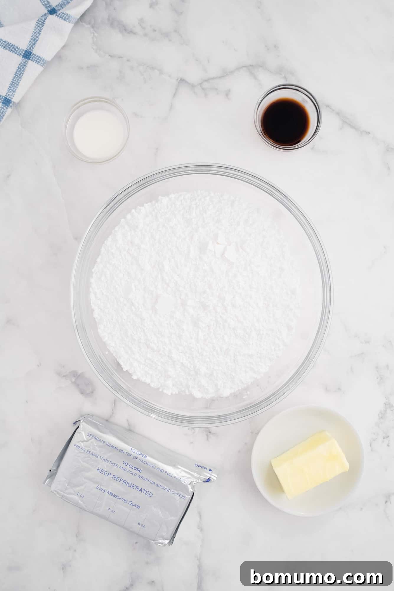 Overhead flat lay of the essential ingredients for cream cheese frosting: softened butter, a block of cream cheese, powdered sugar, vanilla extract, and a small jug of milk.