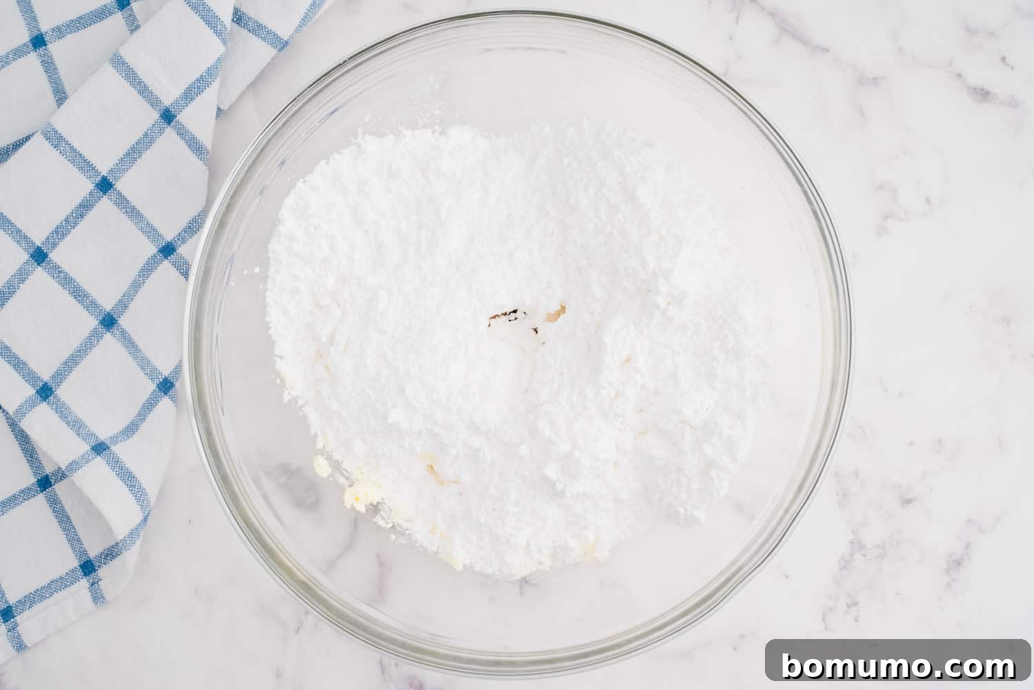 A mixing bowl containing the creamy butter and cream cheese base, with powdered sugar, vanilla extract, and milk added on top, ready for blending.