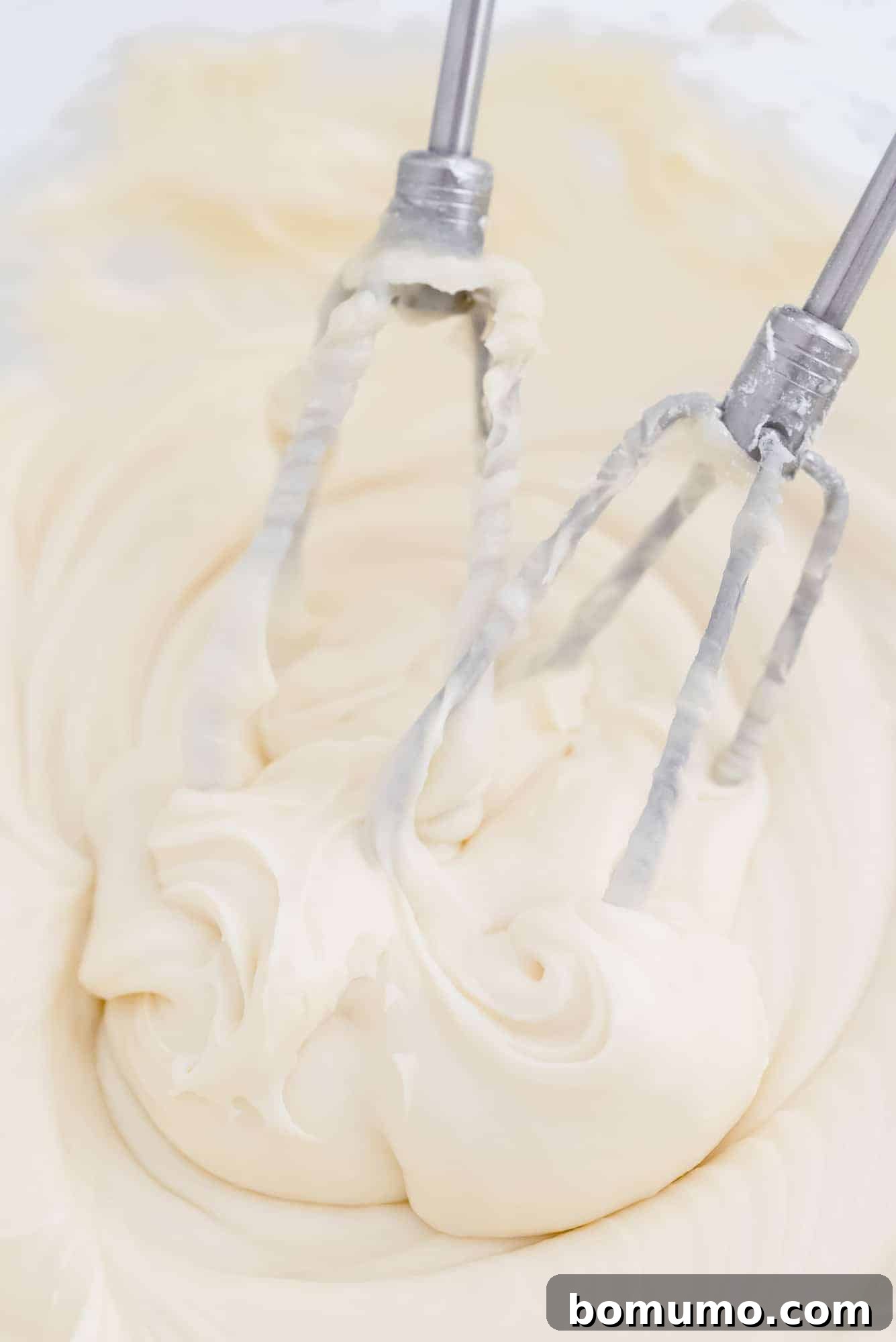 Electric mixer beaters coated in luscious white cream cheese frosting, resting in the bowl after mixing.