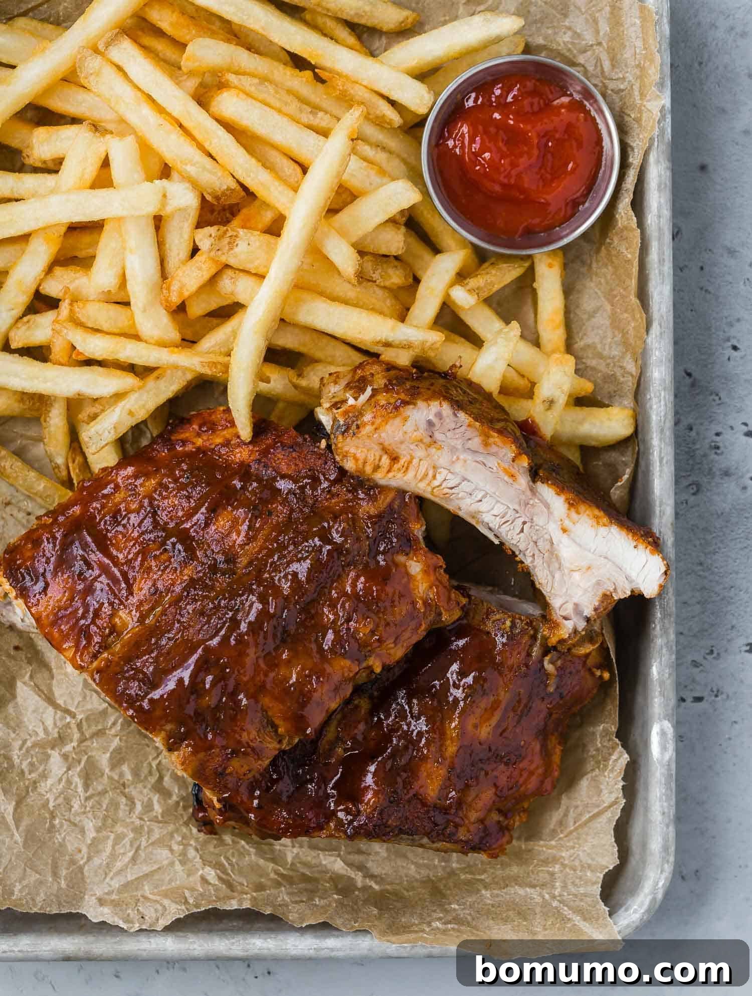 Perfectly cooked ribs on a parchment paper lined tray, served alongside crispy French fries and a small metal cup of ketchup.