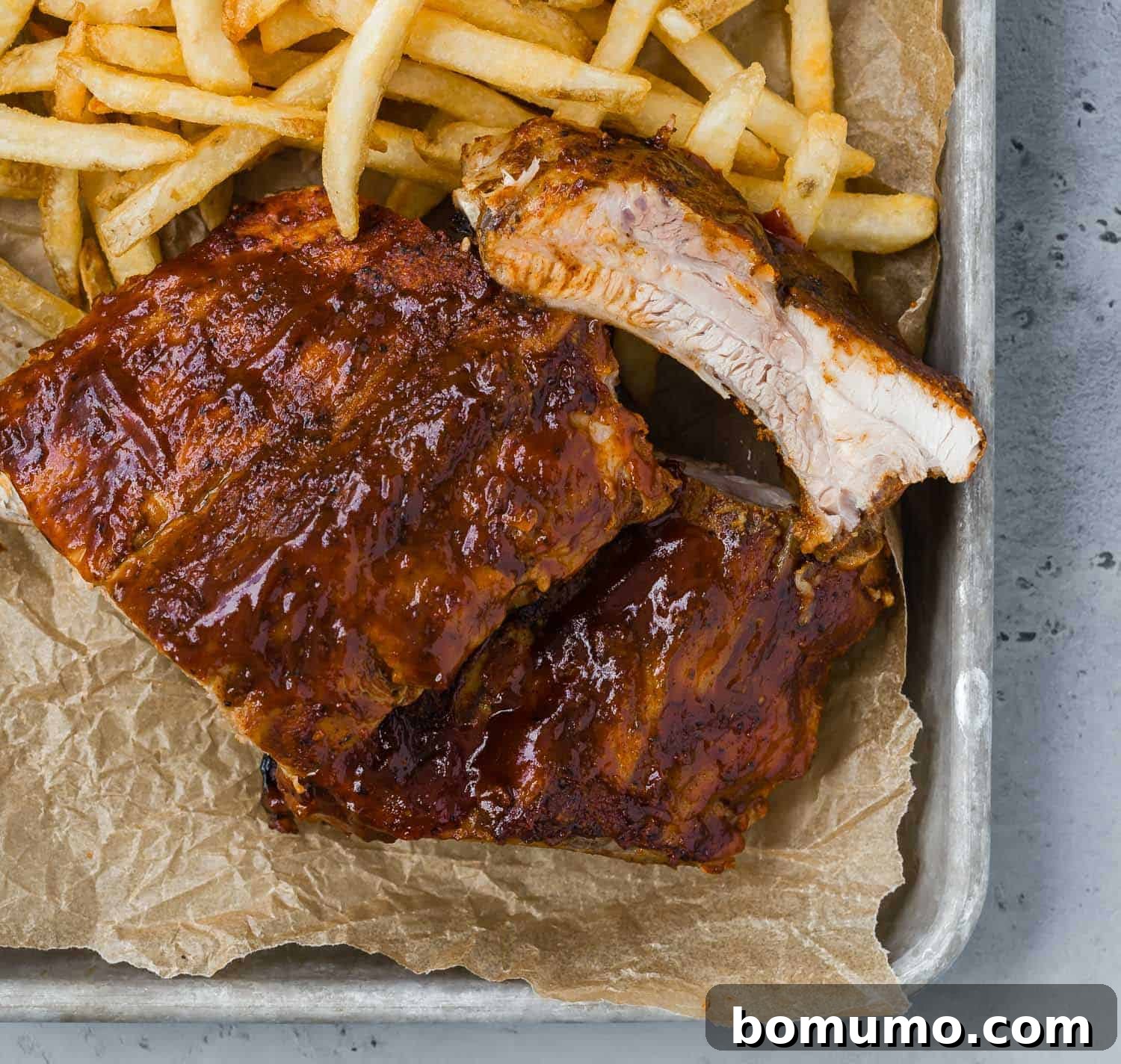A tray lined with brown parchment paper, holding a serving of perfectly cooked BBQ ribs and a generous portion of golden french fries.